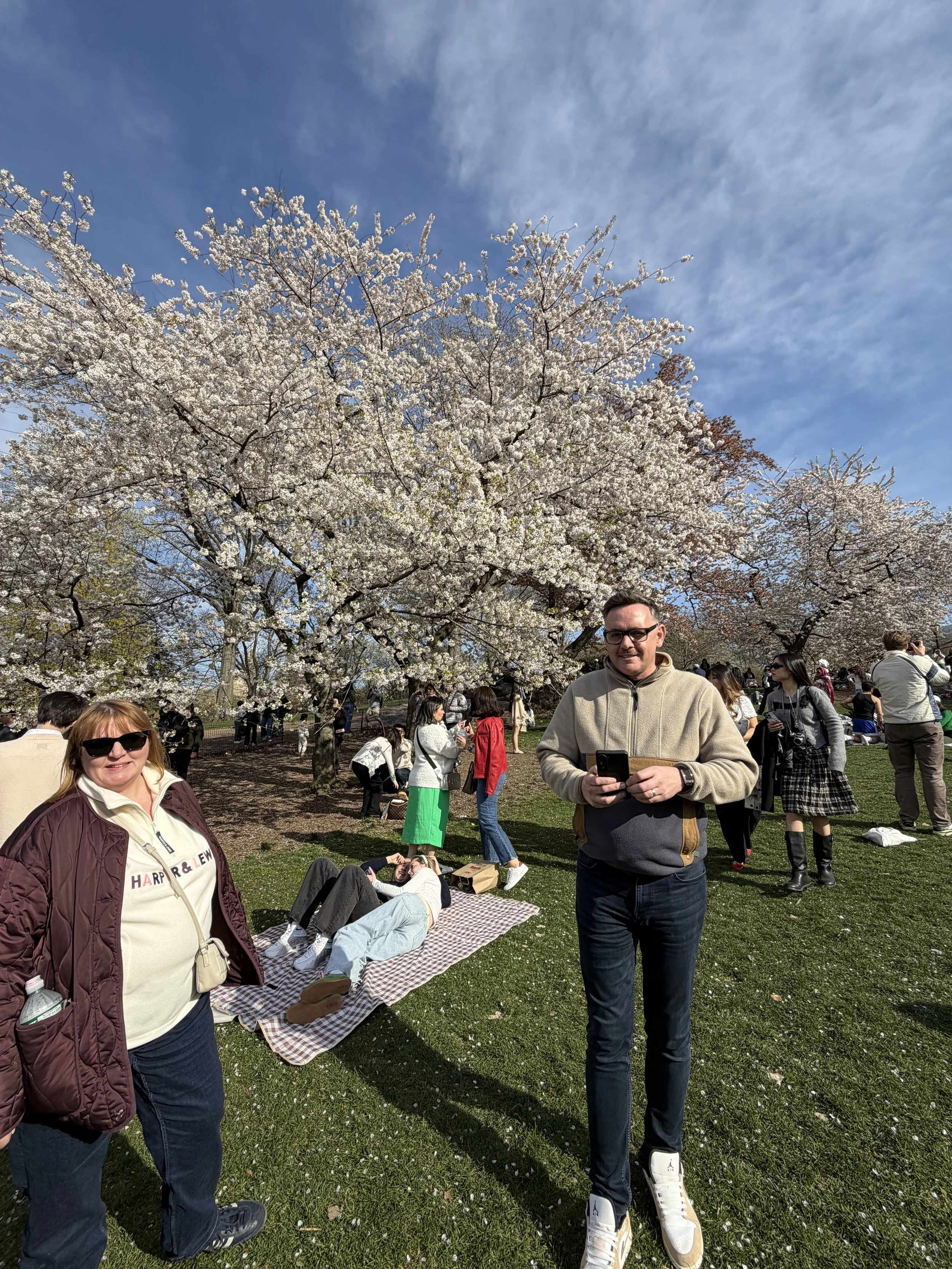 People enjoying a spring day outdoors under blooming cherry blossom trees with blue skies and scattered clouds.