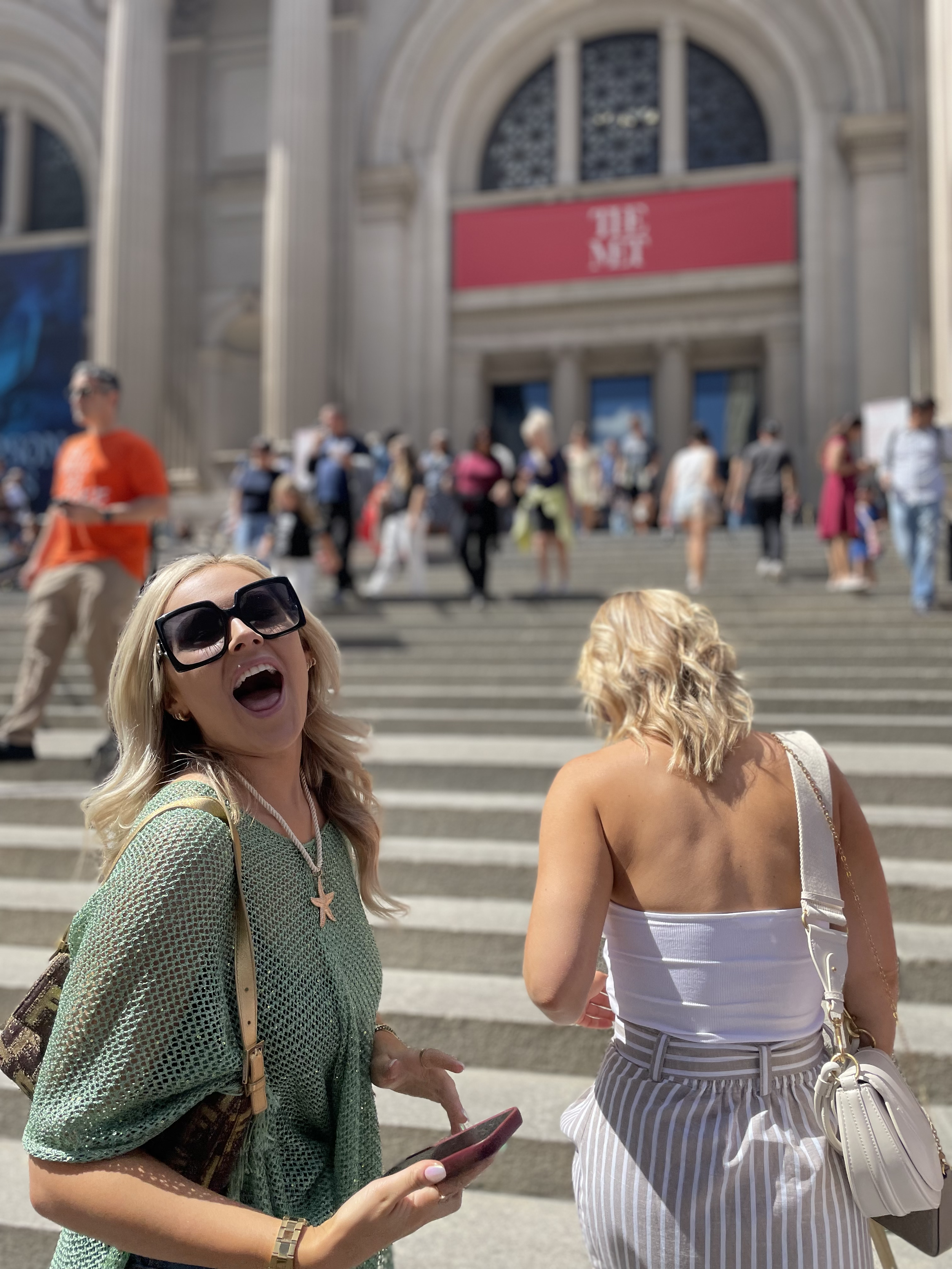 Two women standing on steps outside a large building with a crowd in the background. One woman is smiling, wearing sunglasses, a green mesh top, and holding a phone, and the other woman is with her back to the camera, wearing a white tank top and str