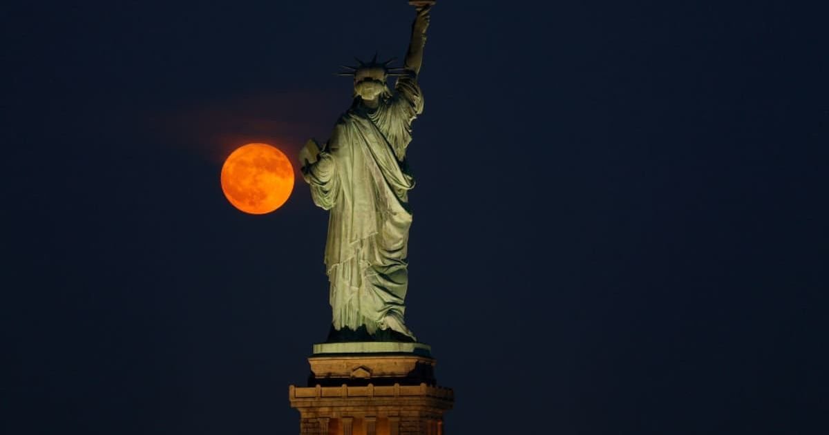Nighttime view of the Statue of Liberty with a full orange moon in the background.