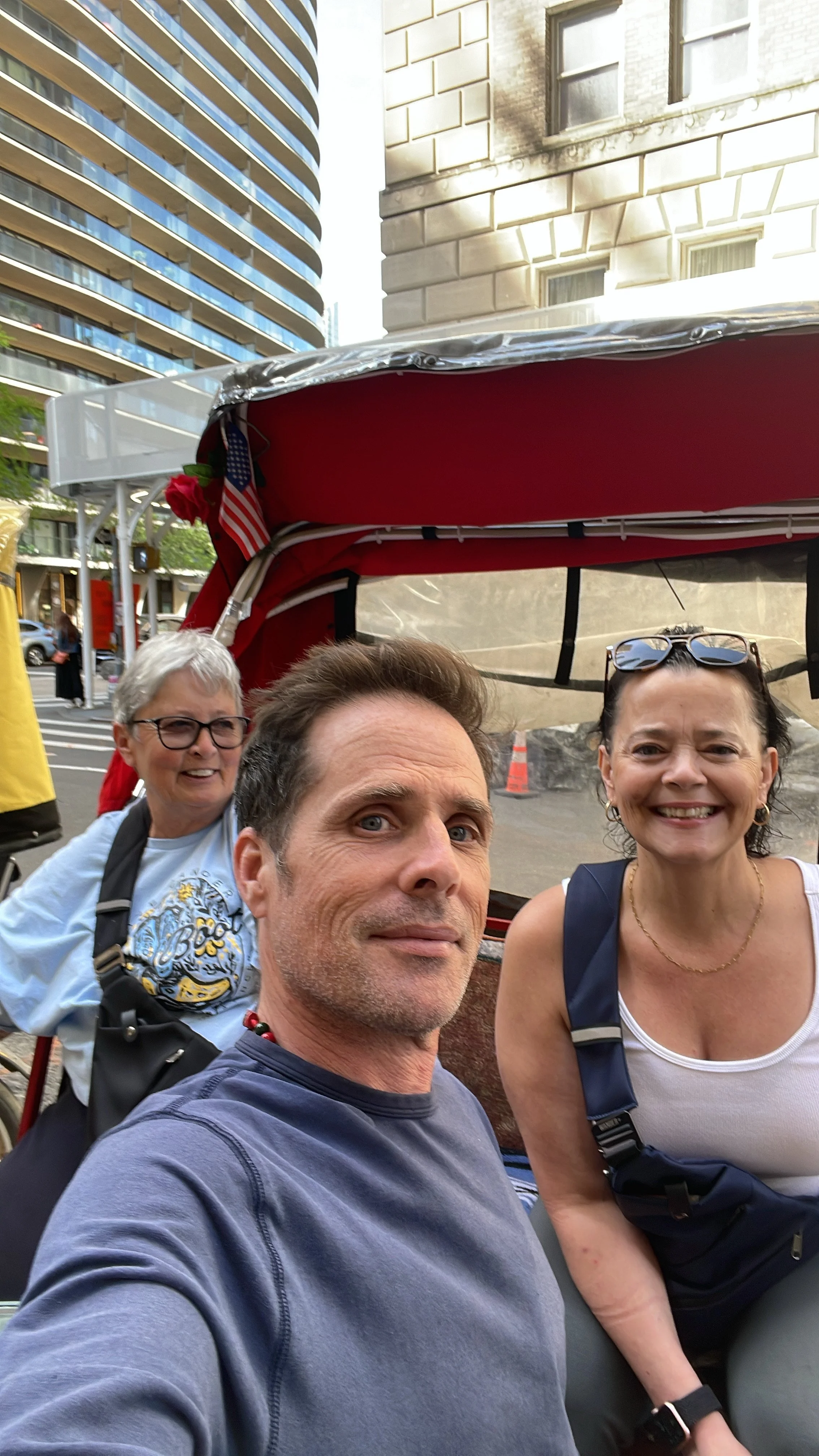 Three smiling people taking a selfie on a city street, with tall buildings in the background.