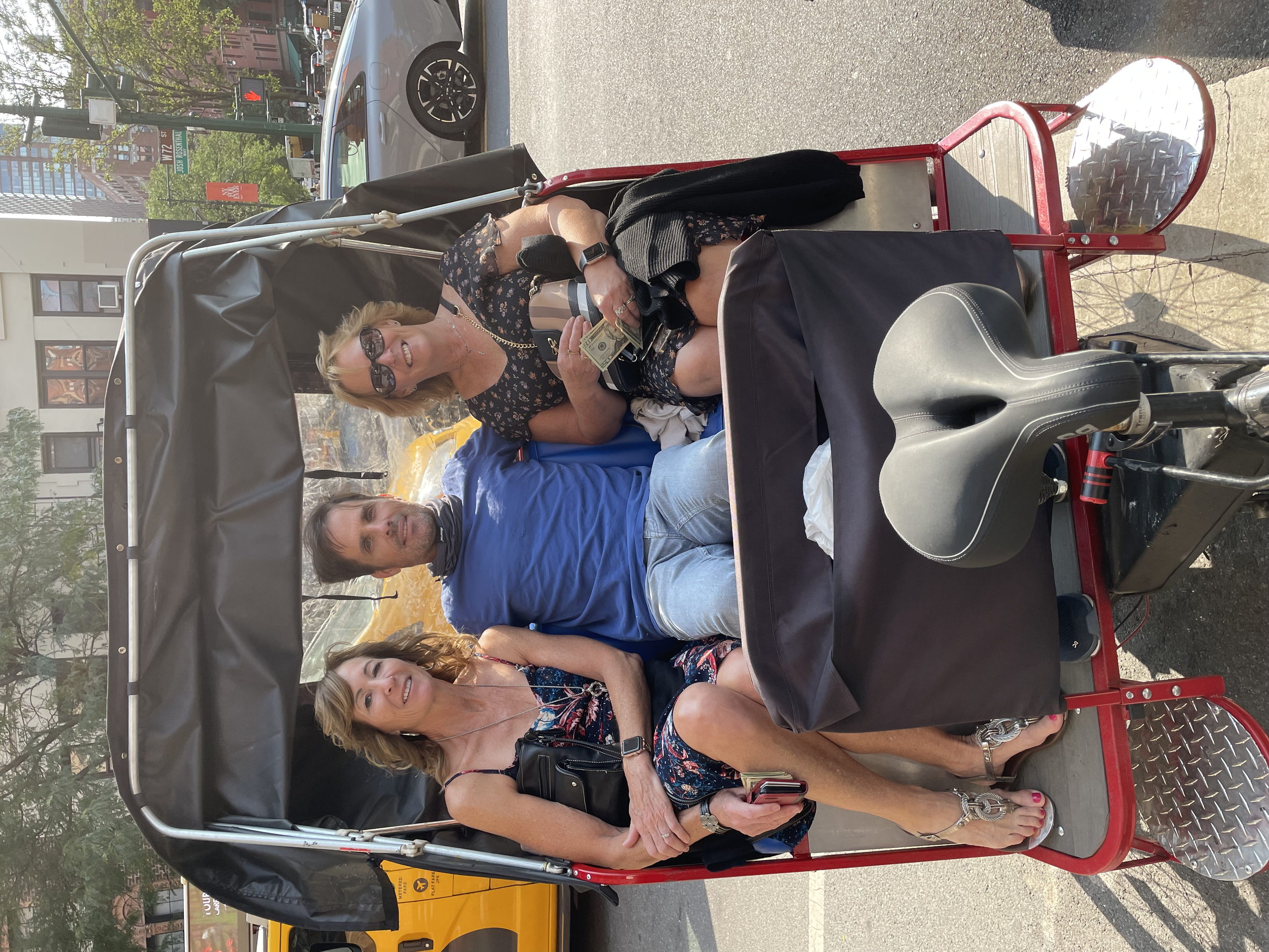 Group of five adults seated in a red open-air tour tram on city street, cityscape background with buildings, trees, and parked cars, the woman in floral top holding dollar bills and smiling.