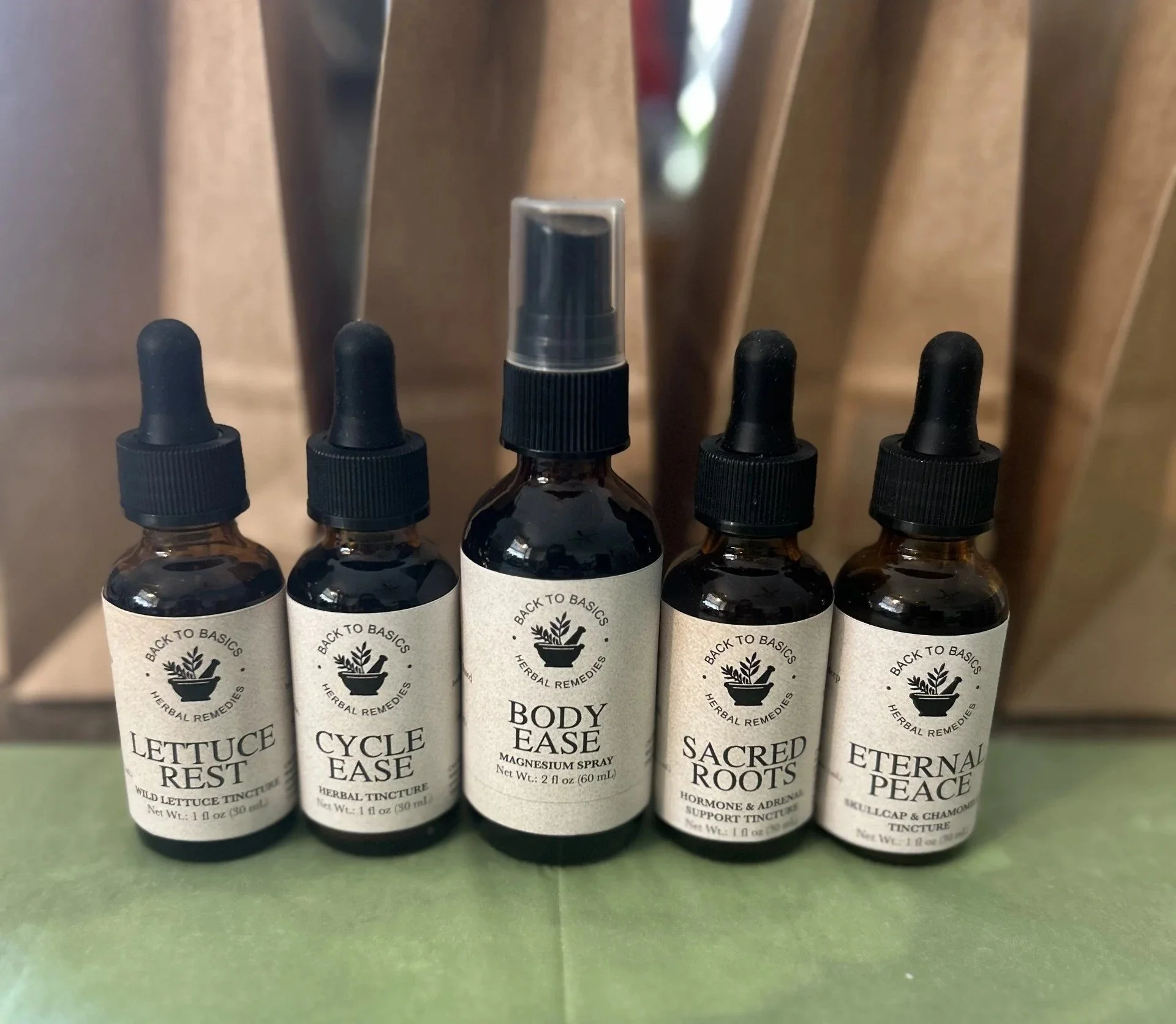 Five small herbal remedy bottles with black dropper caps, labeled from left to right: Lettuce Rest, Cycle Ease, Body Ease, Sacred Roots, Eternal Peace, placed on a green surface with brown bags in the background.
