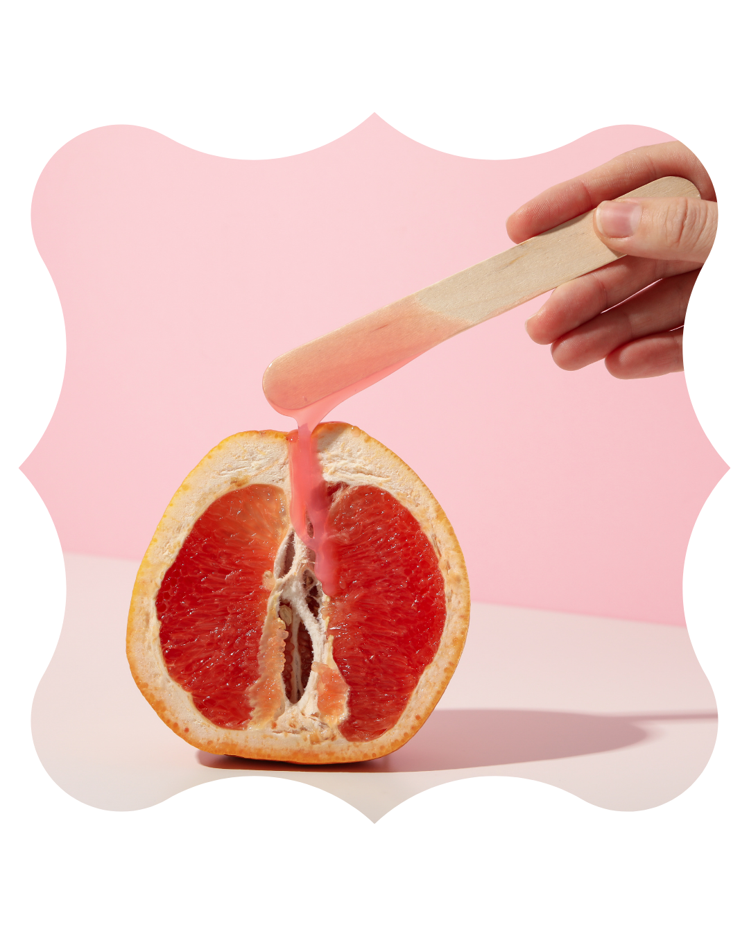 A hand holding a wooden popsicle stick dripping pink wax over a halved pink grapefruit against a pink background.