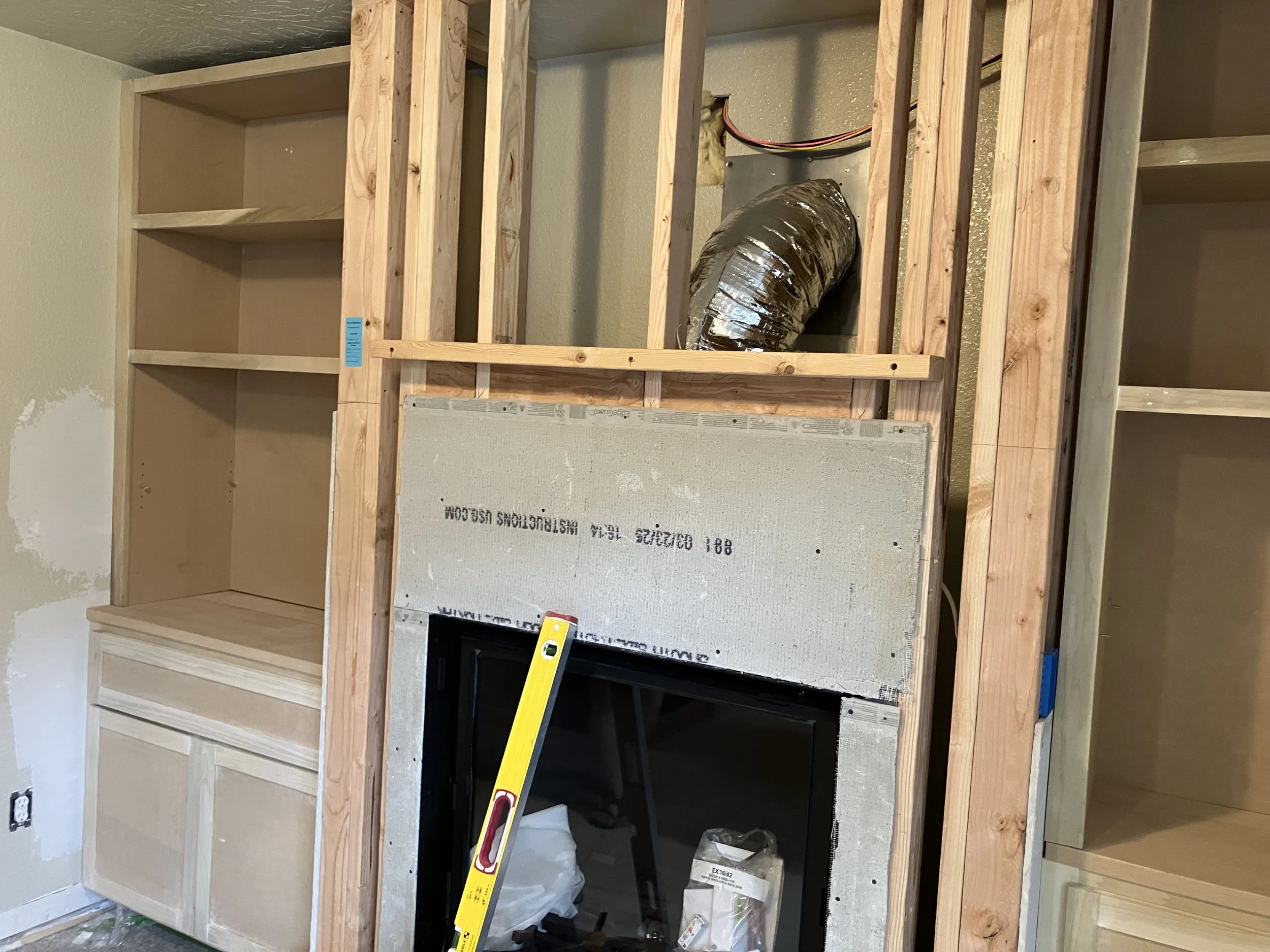 Under construction built-in wooden cabinetry surrounding a fireplace with a vent pipe above, a yellow level leaning against the fireplace opening, and unfinished walls.