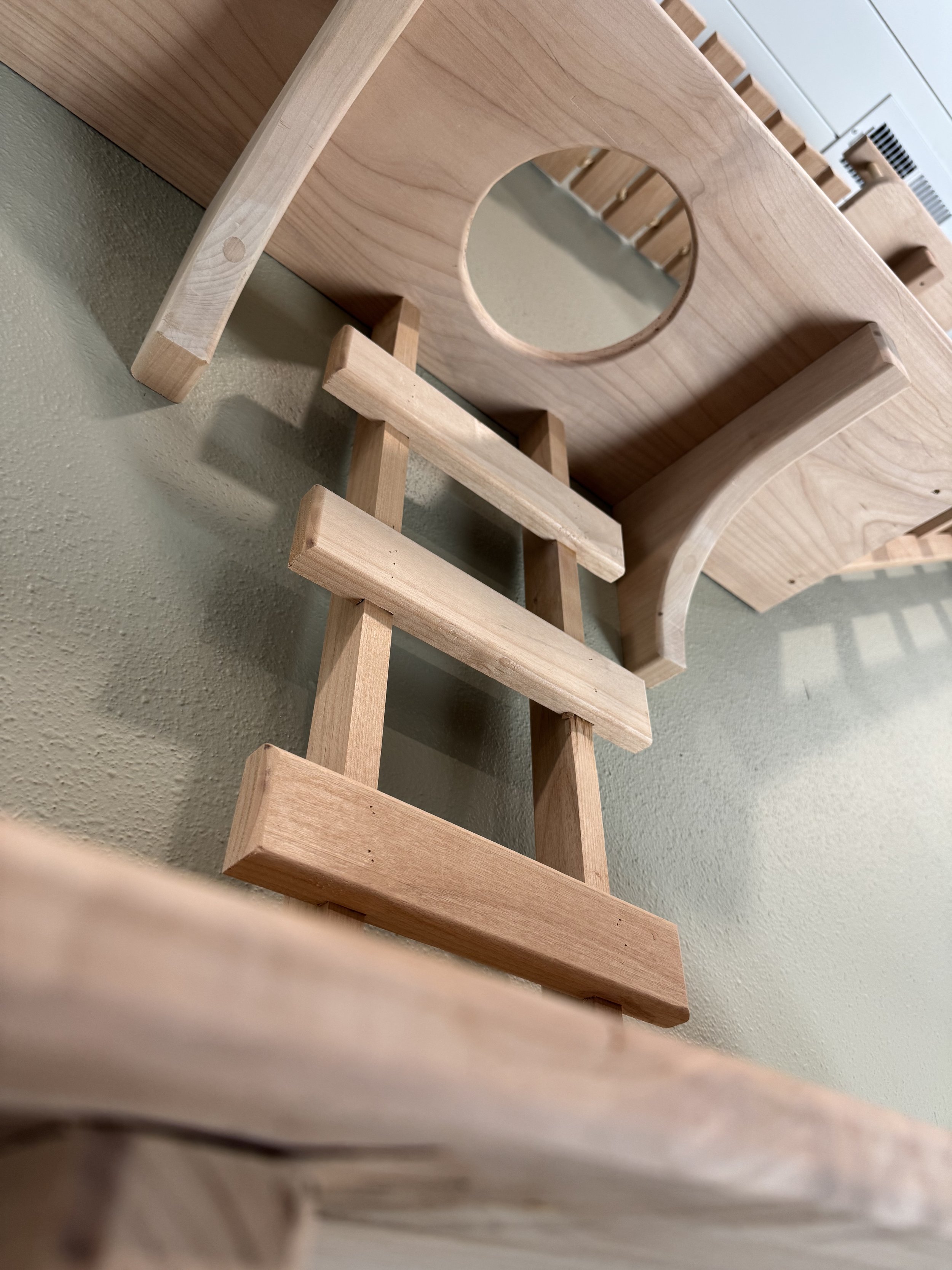 Close-up of unfinished wooden furniture pieces, including a stool and a piece with a circular cutout, against a textured wall and a white cabinet with black vent and wooden slats.