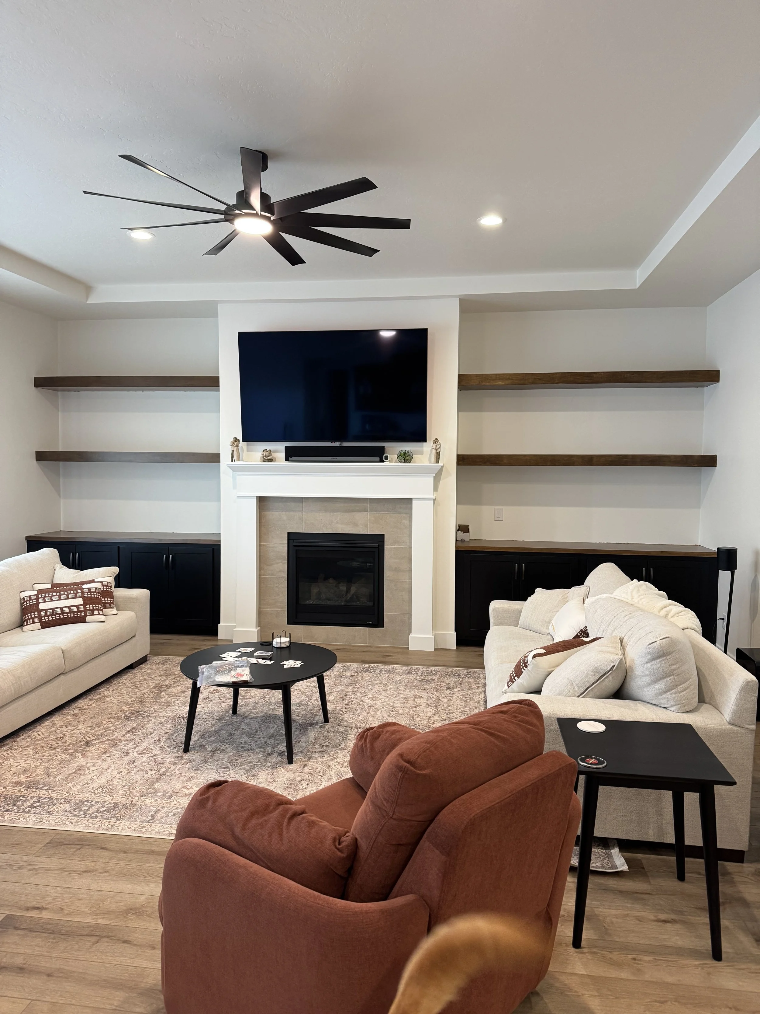 Living room with beige sofas, a rust-colored armchair, a black coffee table, a built-in entertainment center, a wall-mounted TV, a fireplace, and wooden shelving.