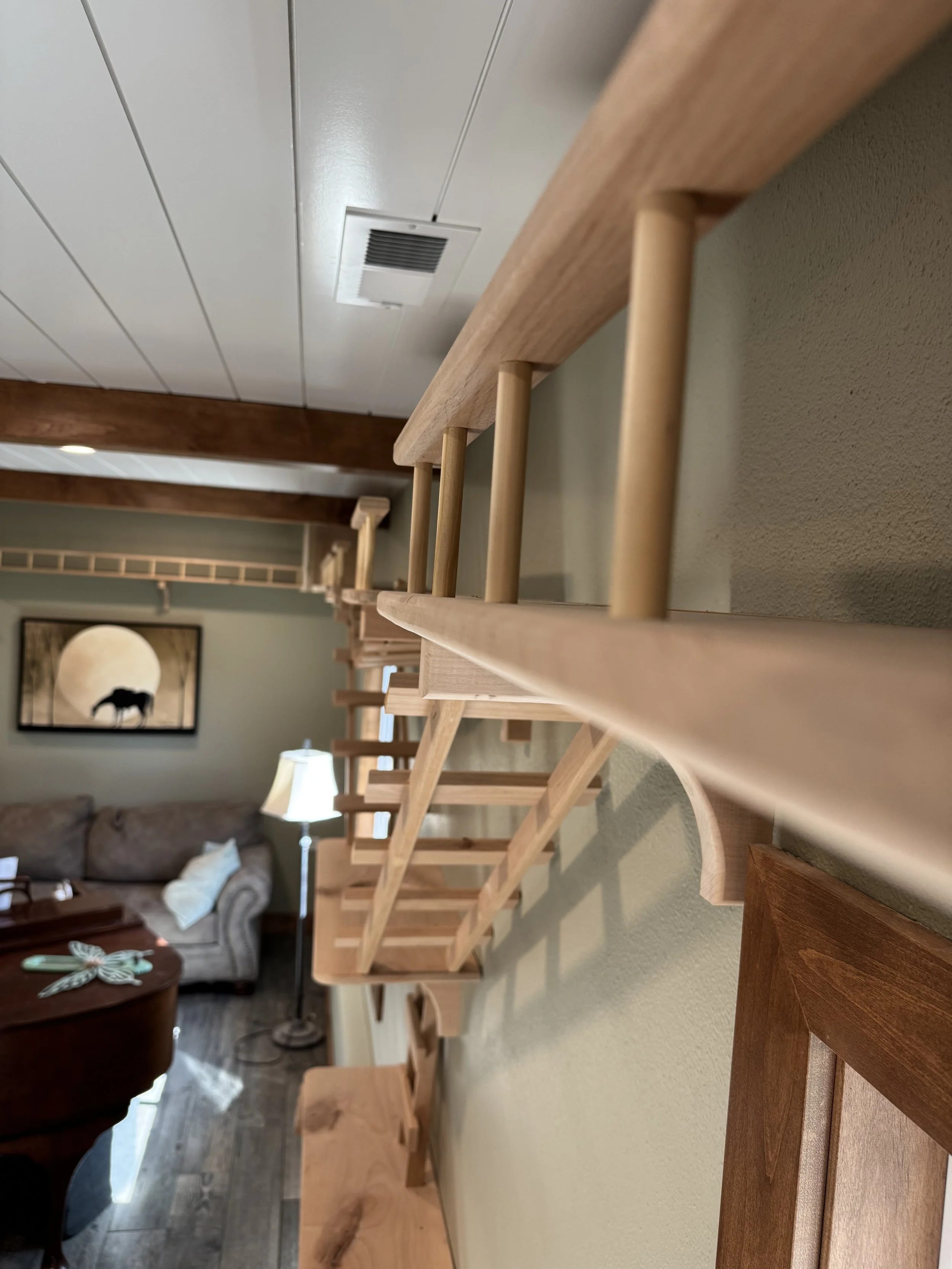 A wooden staircase with vertical spindles and a handrail, viewed from below in a living room with a beige couch, a wall picture of a wolf and moon, and a standing lamp.