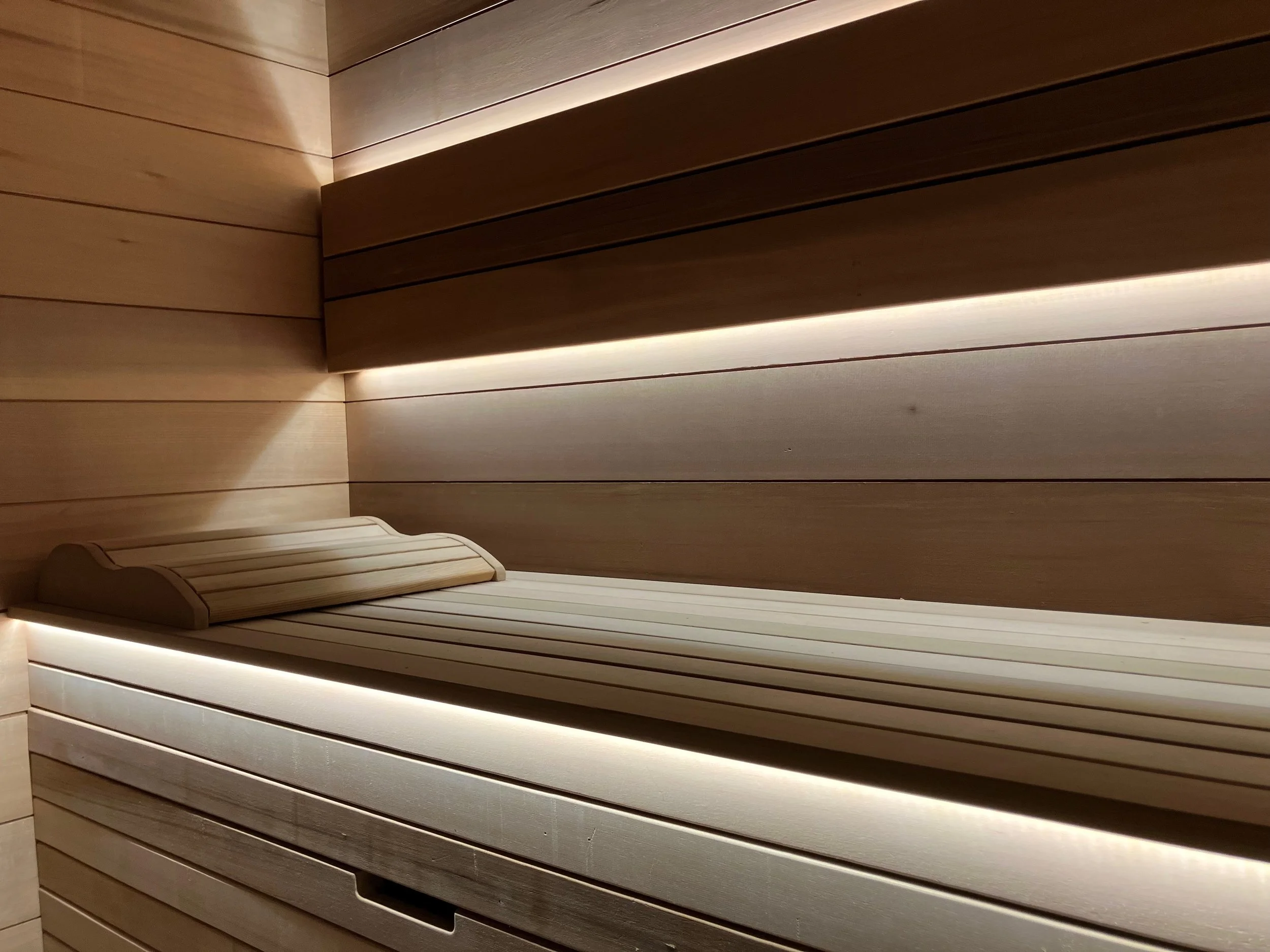 Empty wooden sauna with a wood lounge chair, soft lighting, minimal decor, and horizontal wood paneling.