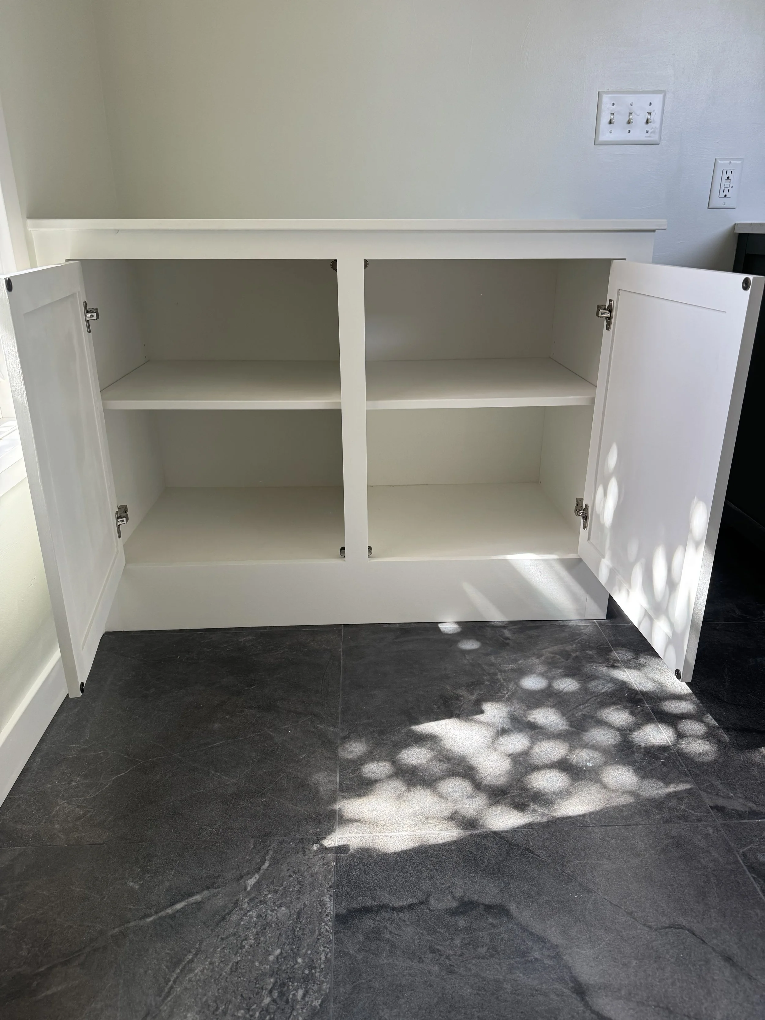 Empty white cabinet with two doors open, situated on dark tiled floor with sunlight and shadow patterns
