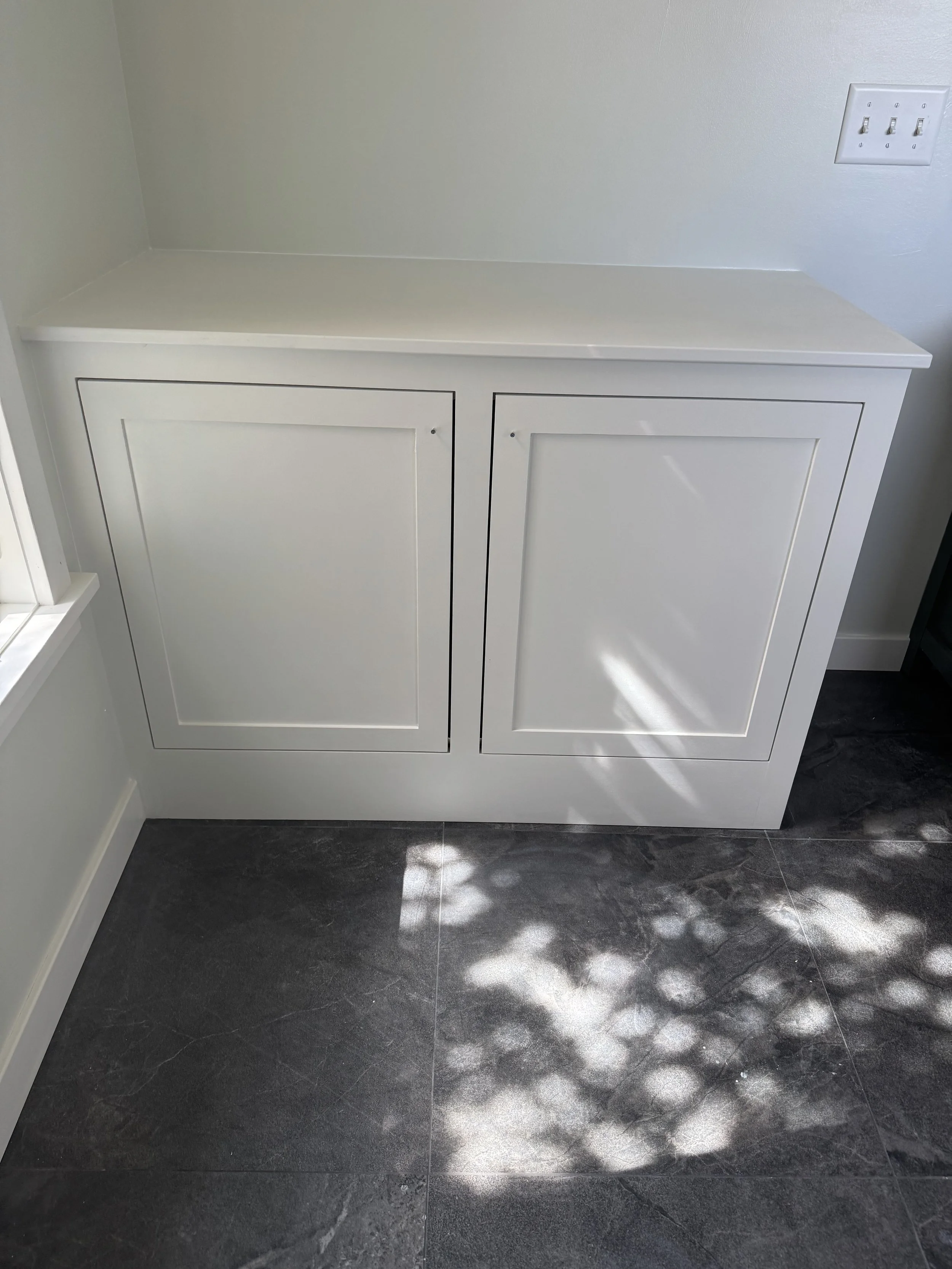 White cabinet with double doors against a white wall, next to a window casting light and shadows on dark tiled floor.