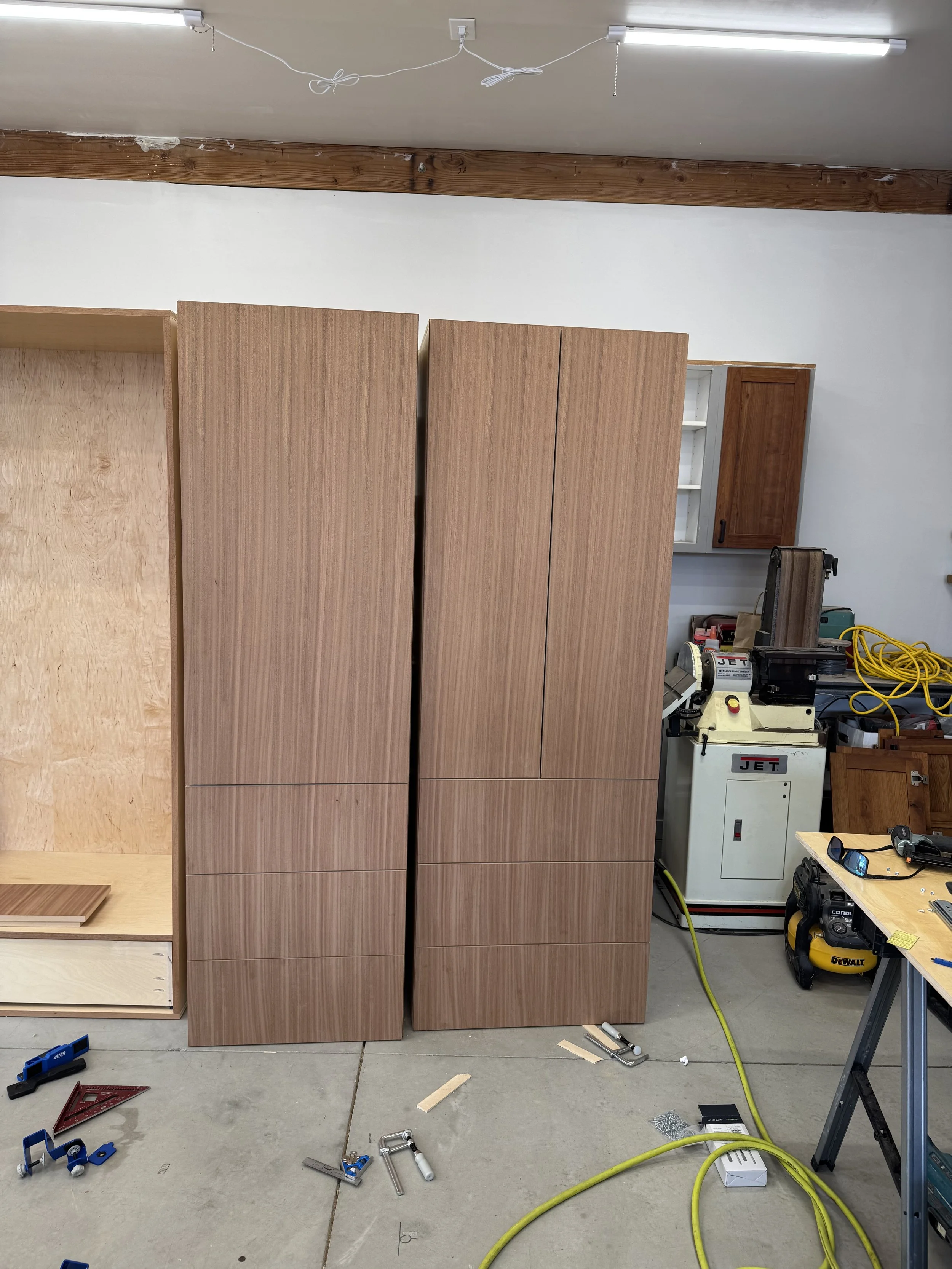 Two tall wooden cabinets in a woodworking workshop, with tools and supplies on a workbench nearby.