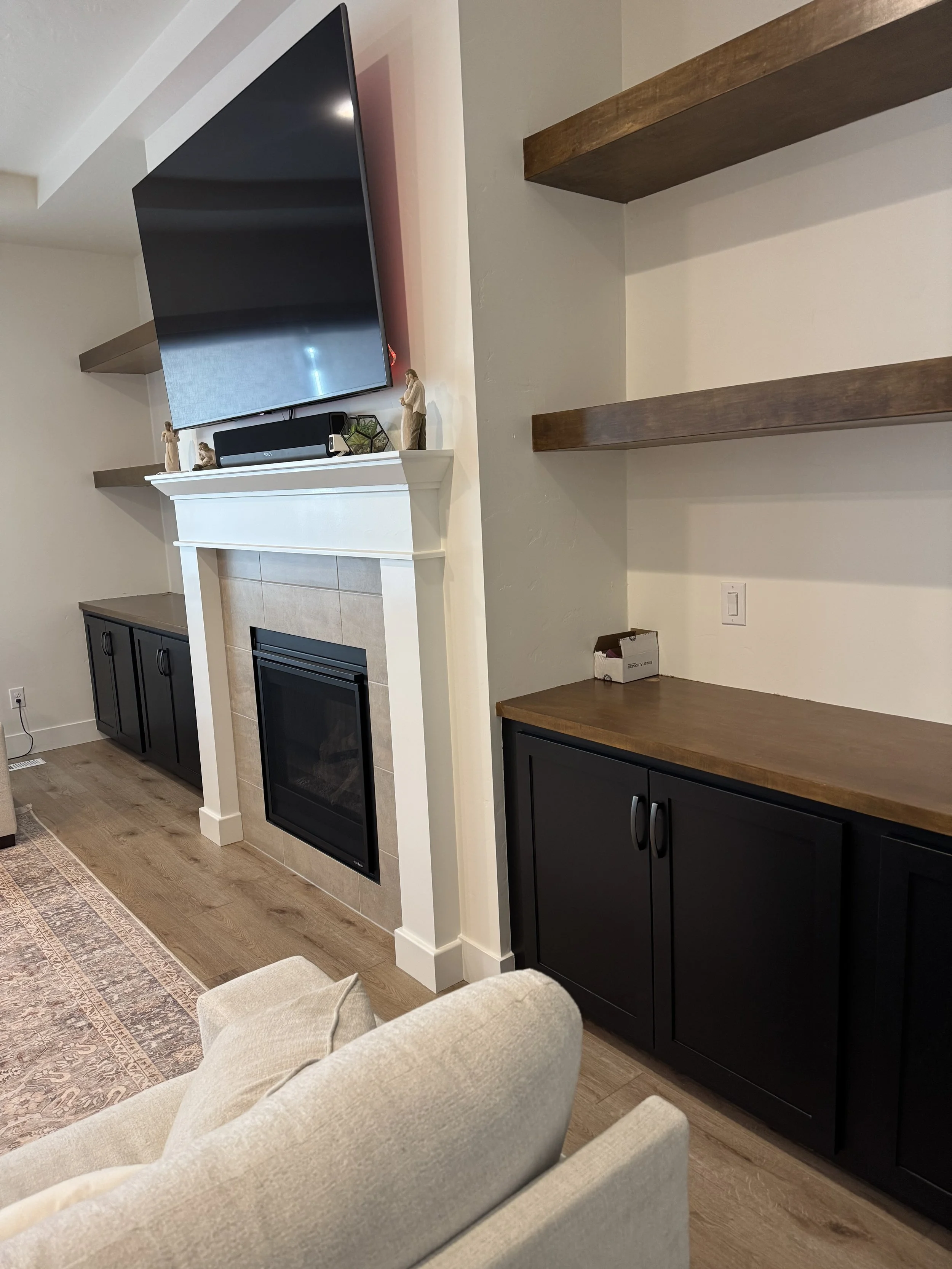 Living room with fireplace, mounted TV, decorative figurines on mantel, black cabinets, wooden shelves on the wall, and beige upholstered furniture.