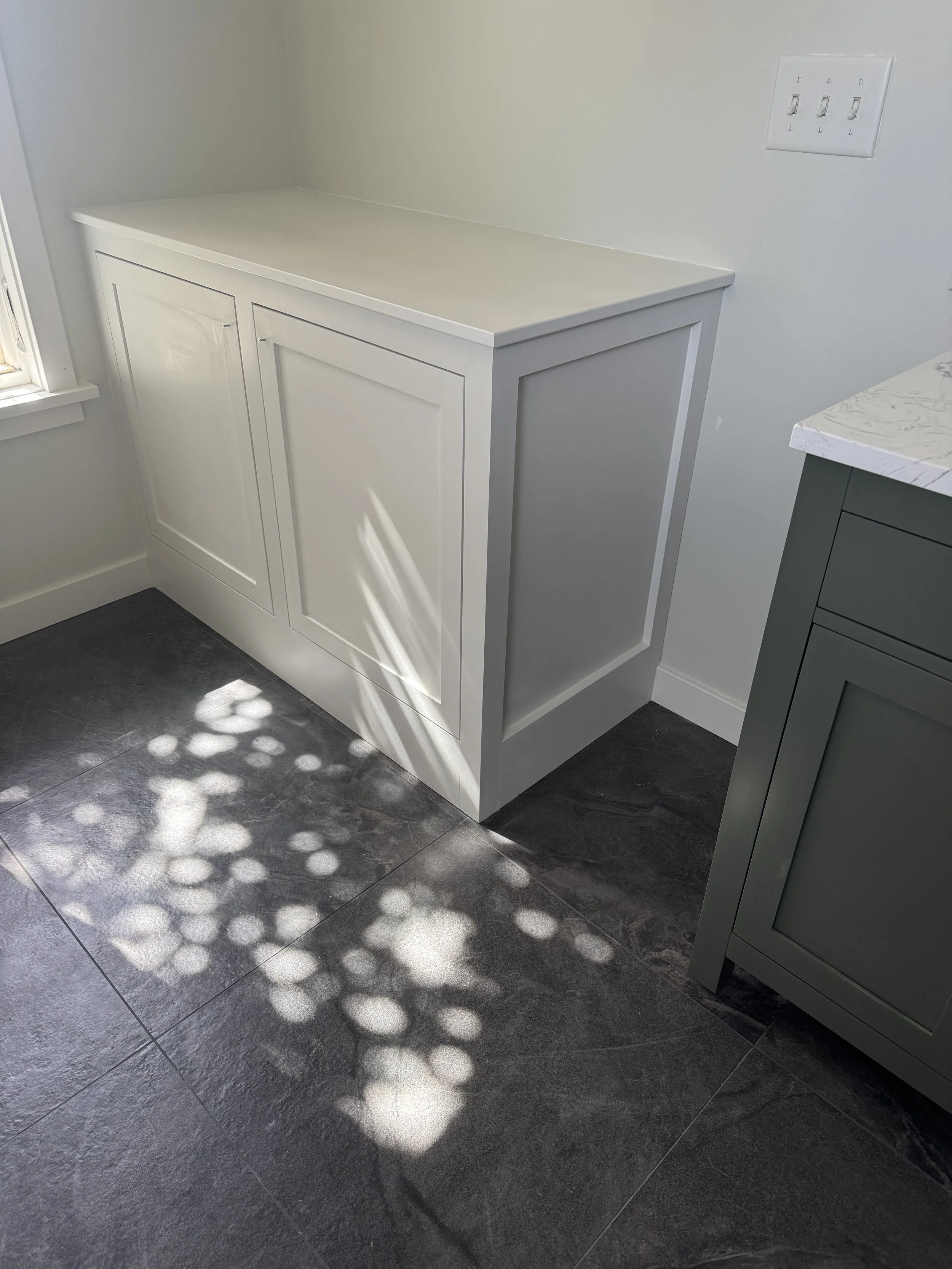 White kitchen cabinet with a white countertop near a window, with shadow of leaves on dark tiled floor.