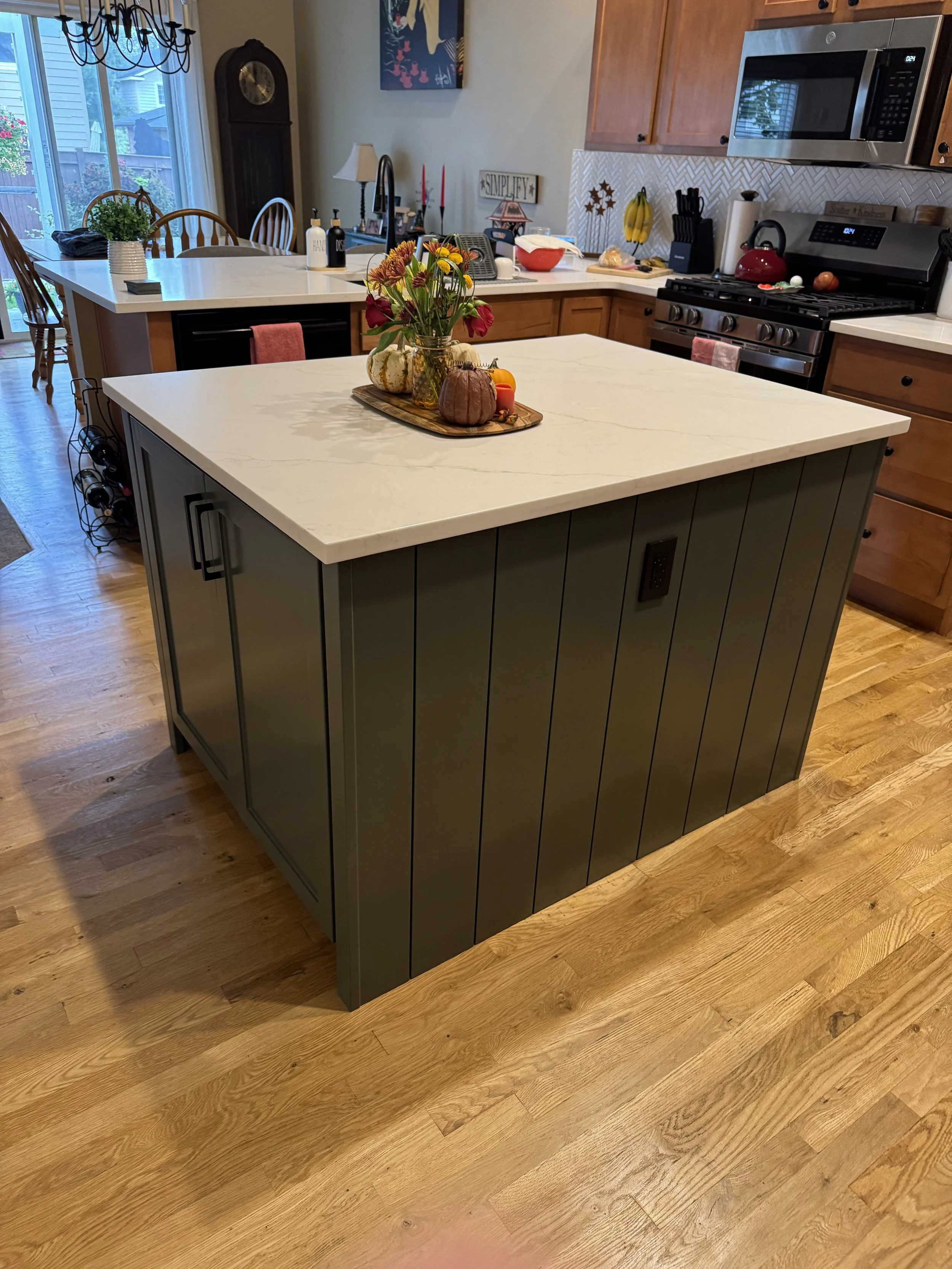 A kitchen island with a white marble top and dark gray paneling on the sides, decorated with a flower arrangement and small pumpkins. In the background, there are wooden cabinets, a stove, microwave, and various kitchen items on the counters.