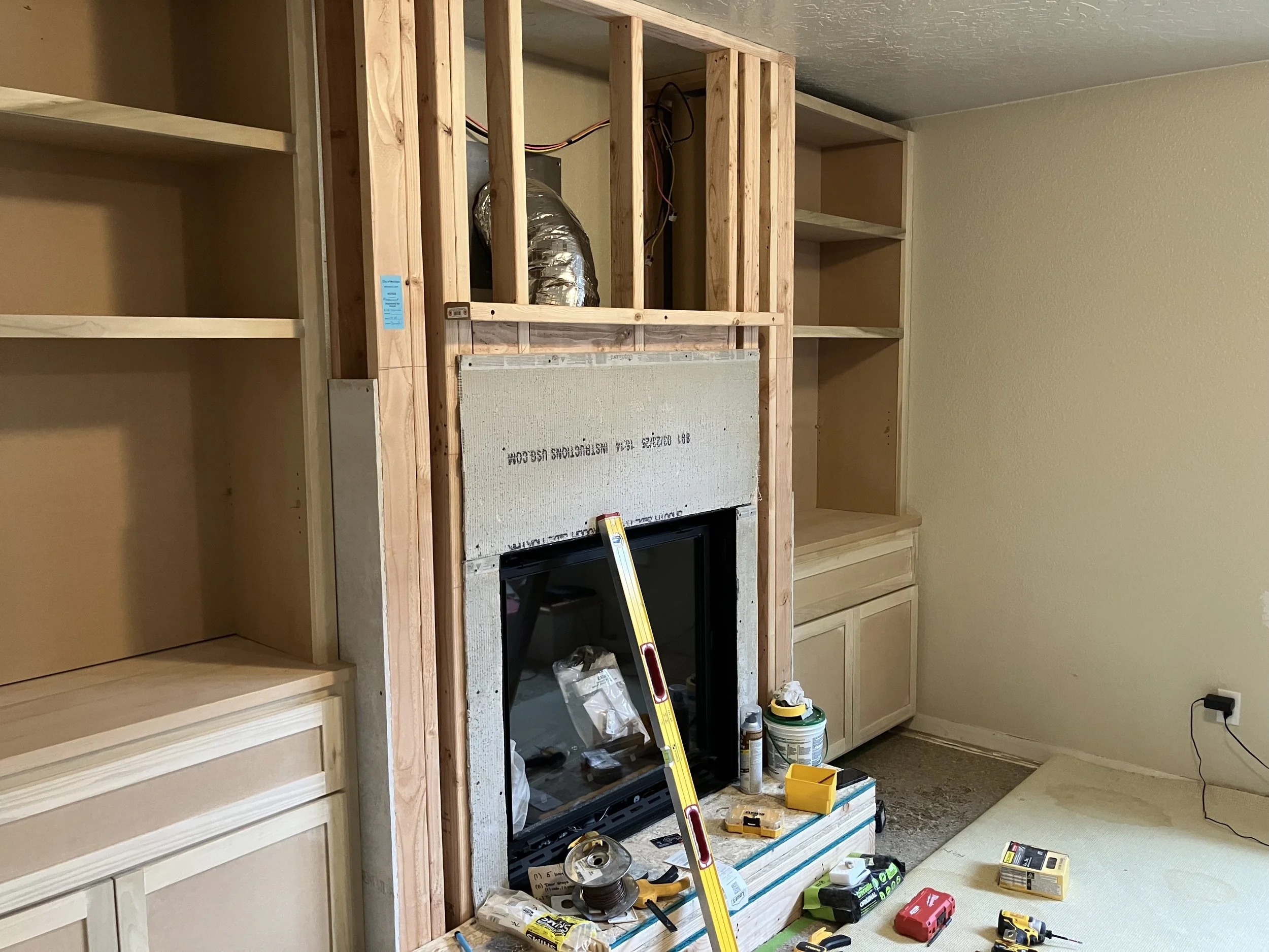 Room under renovation with unfinished wooden framing around a new fireplace installation. Building tools, supplies, and a ladder are scattered on the floor, with empty shelving units on either side.