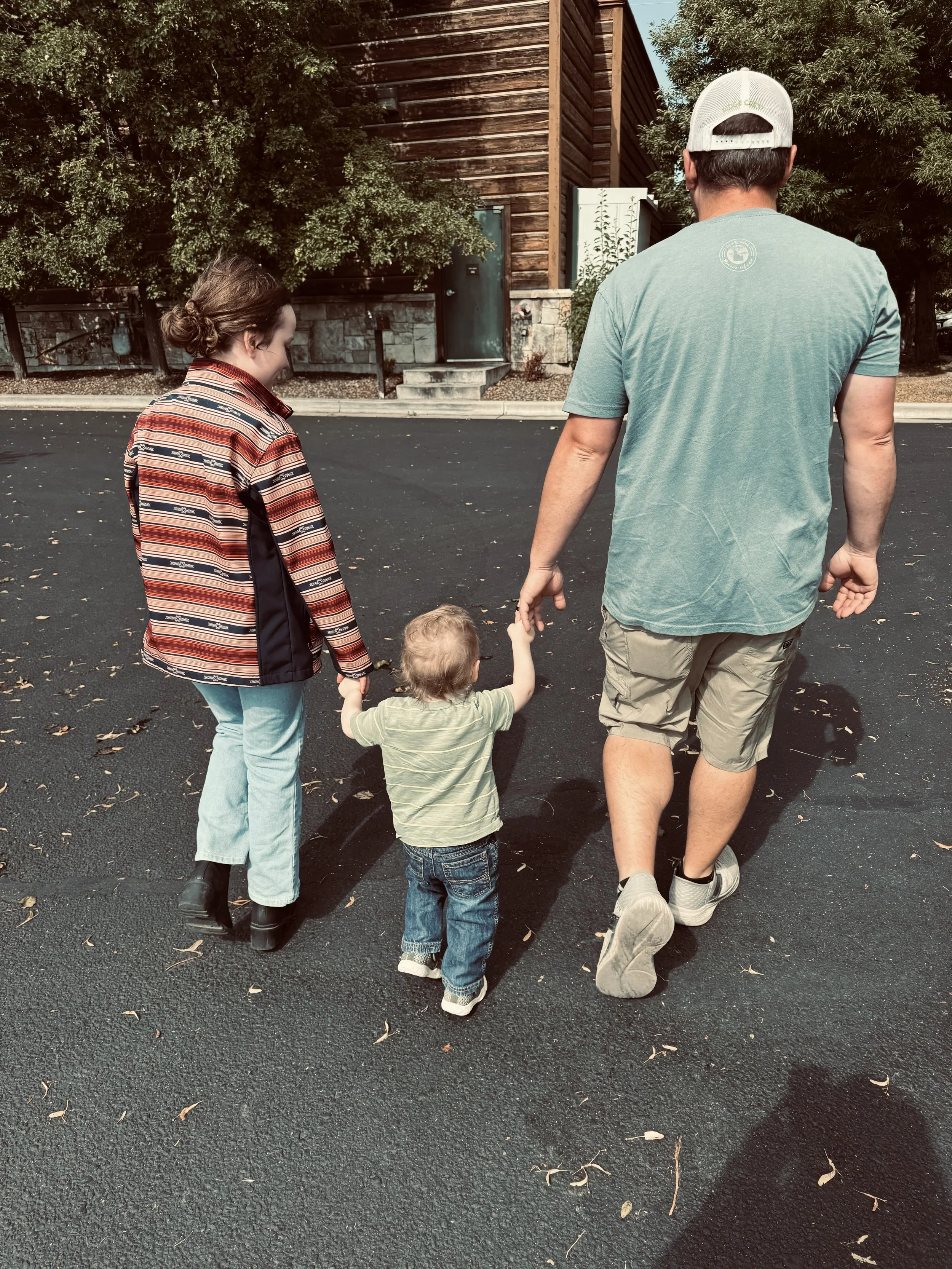 A family walking on a paved street. A woman on the left wearing a patterned jacket and light jeans is holding the hand of a small child in the middle wearing a green striped shirt and jeans. A man on the right in a teal t-shirt, khaki shorts, and a cap is holding the child's other hand. There are trees and a wooden building in the background.
