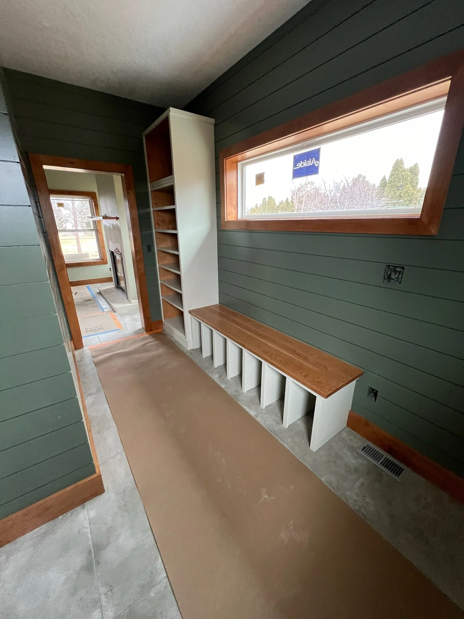A hallway with green wooden walls, a large window, a built-in wooden bench, and a white bookshelf. The floor is covered with a brown protective sheet. Through the doorway, part of a room with a fireplace and another window is visible.