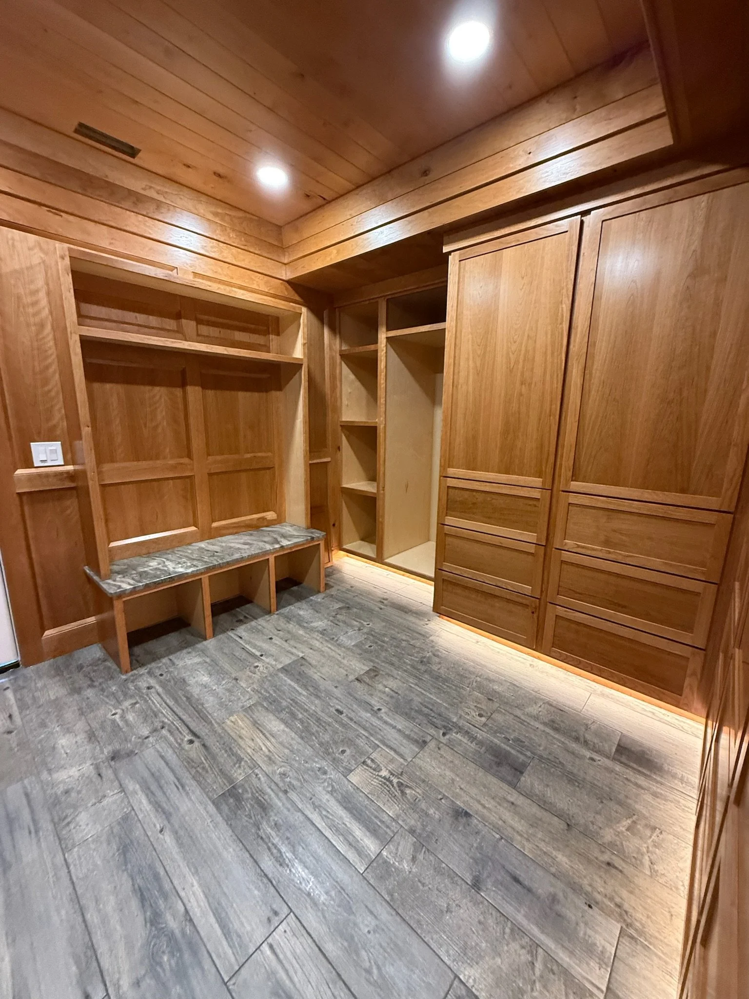 An empty wooden walk-in closet with built-in shelves, drawers, and a bench with a marble top. The closet has wood-paneled walls and ceiling, with recessed ceiling lights and LED lighting along the base of the cabinets.