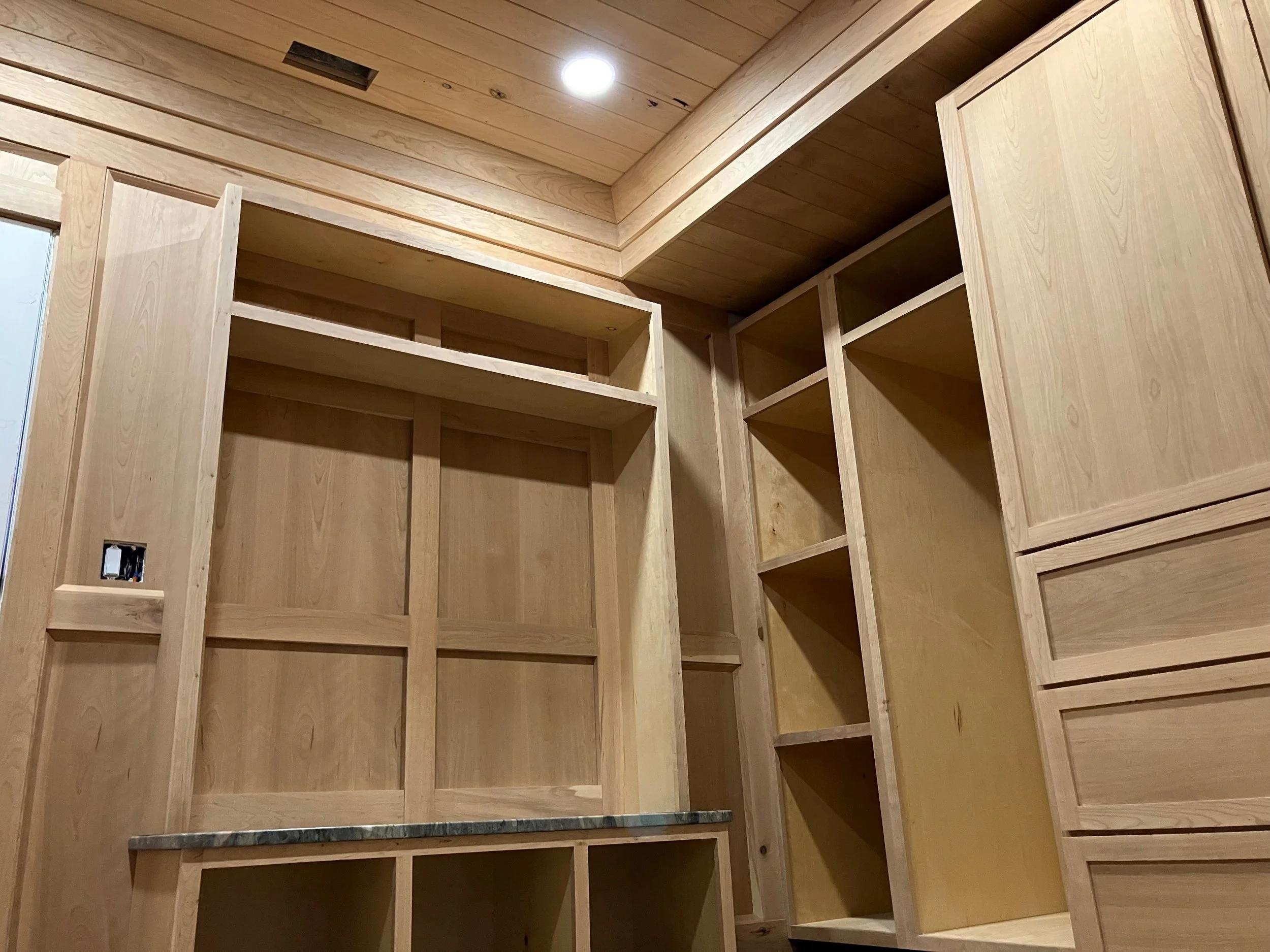 Unfinished wooden closet with multiple shelves and drawers in a room with a wooden ceiling and a ceiling light.
