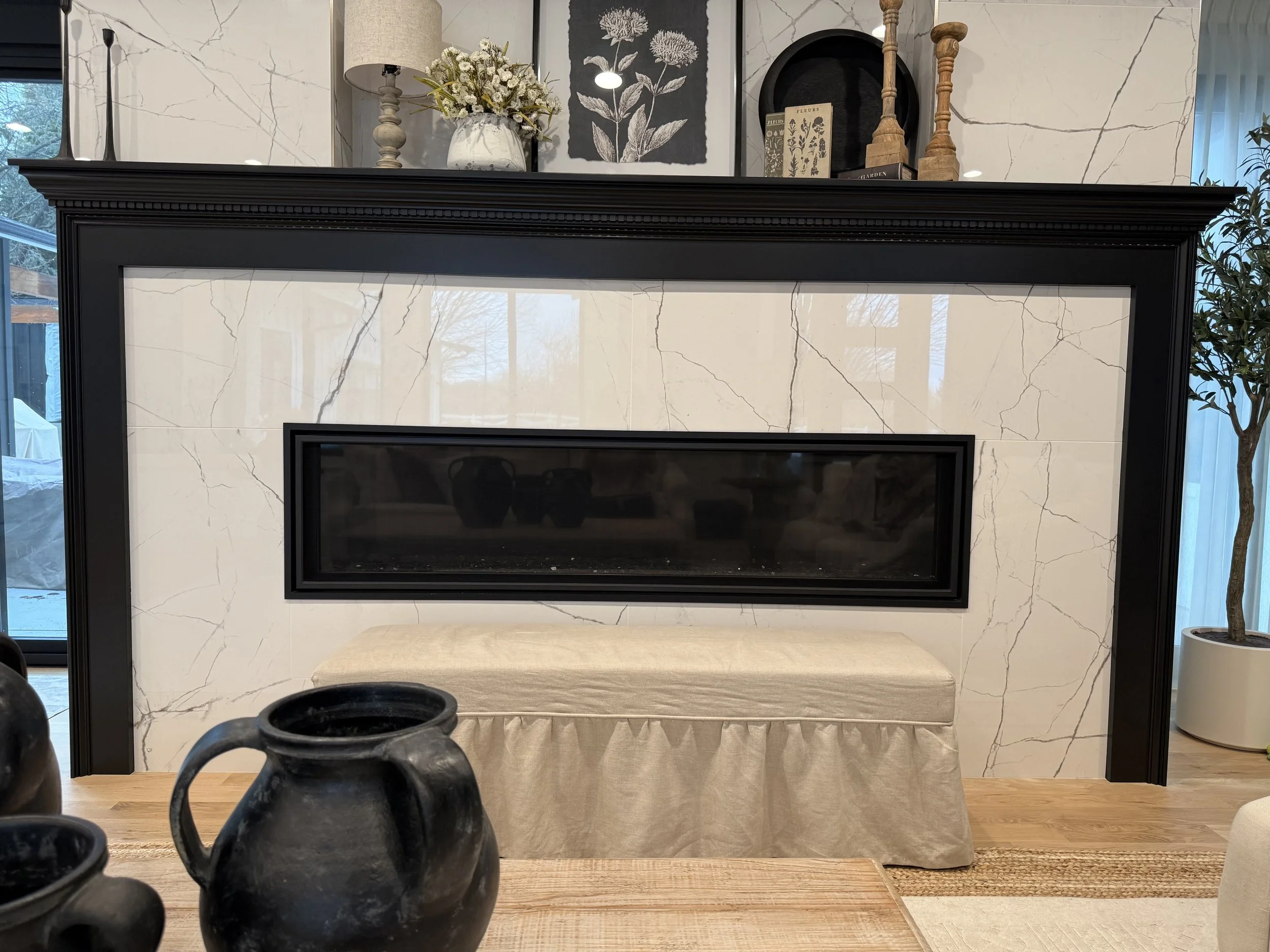 Living room with a fireplace framed in black, decorated with floral artwork, vases, and candles on mantel, beige cushioned bench in front, black vases in foreground, and potted plant near window.