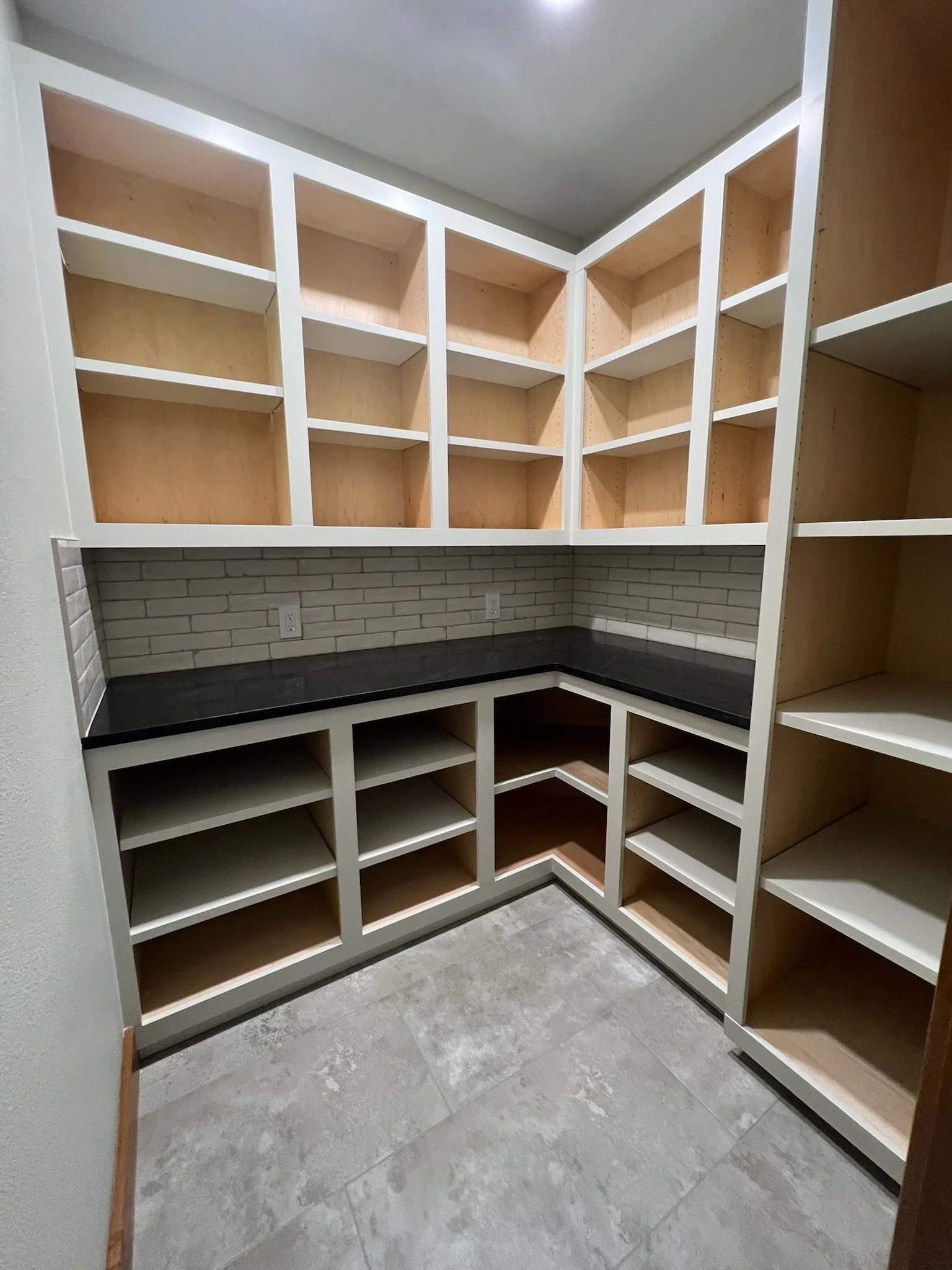 Empty corner pantry with multiple wood shelves, a black countertop, brick wall backsplash, and gray tiled floor.