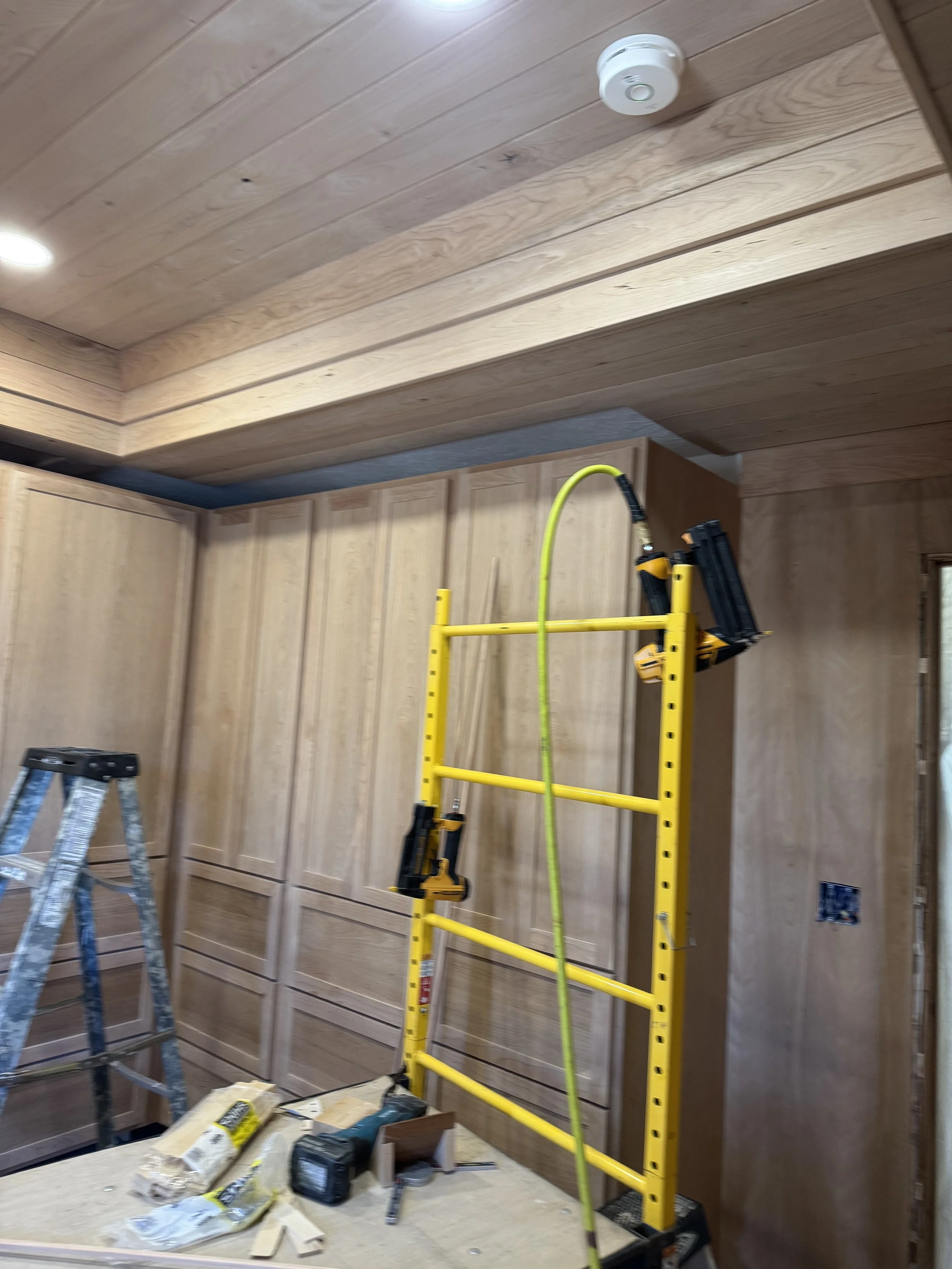 Interior of a room under construction with wood paneling, a yellow scaffolding, a metal step ladder, and construction tools on a work surface.