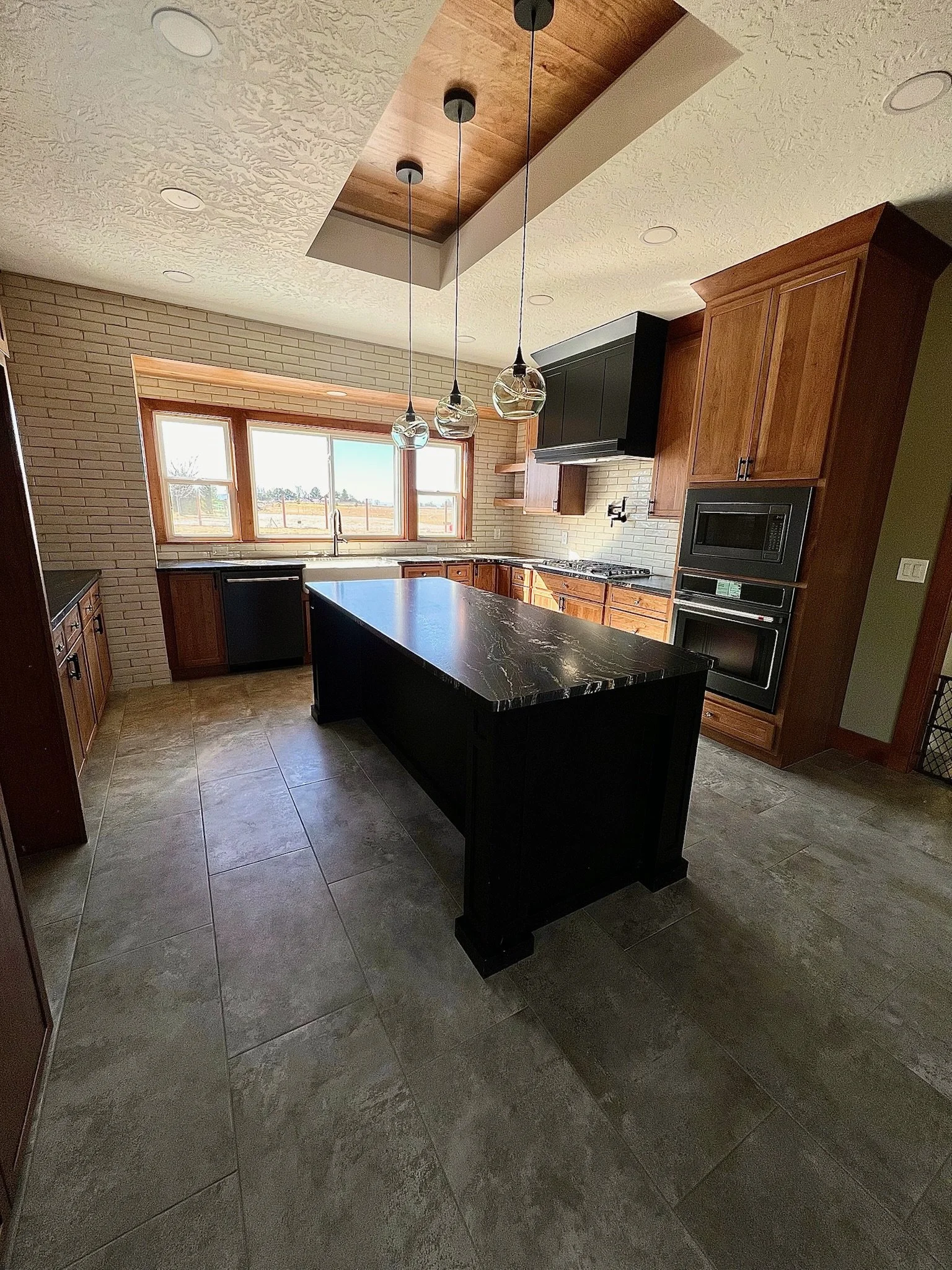 Modern kitchen with large window, wooden cabinets, black appliances, and a black kitchen island with a marble top, pendant lights, and tile flooring.