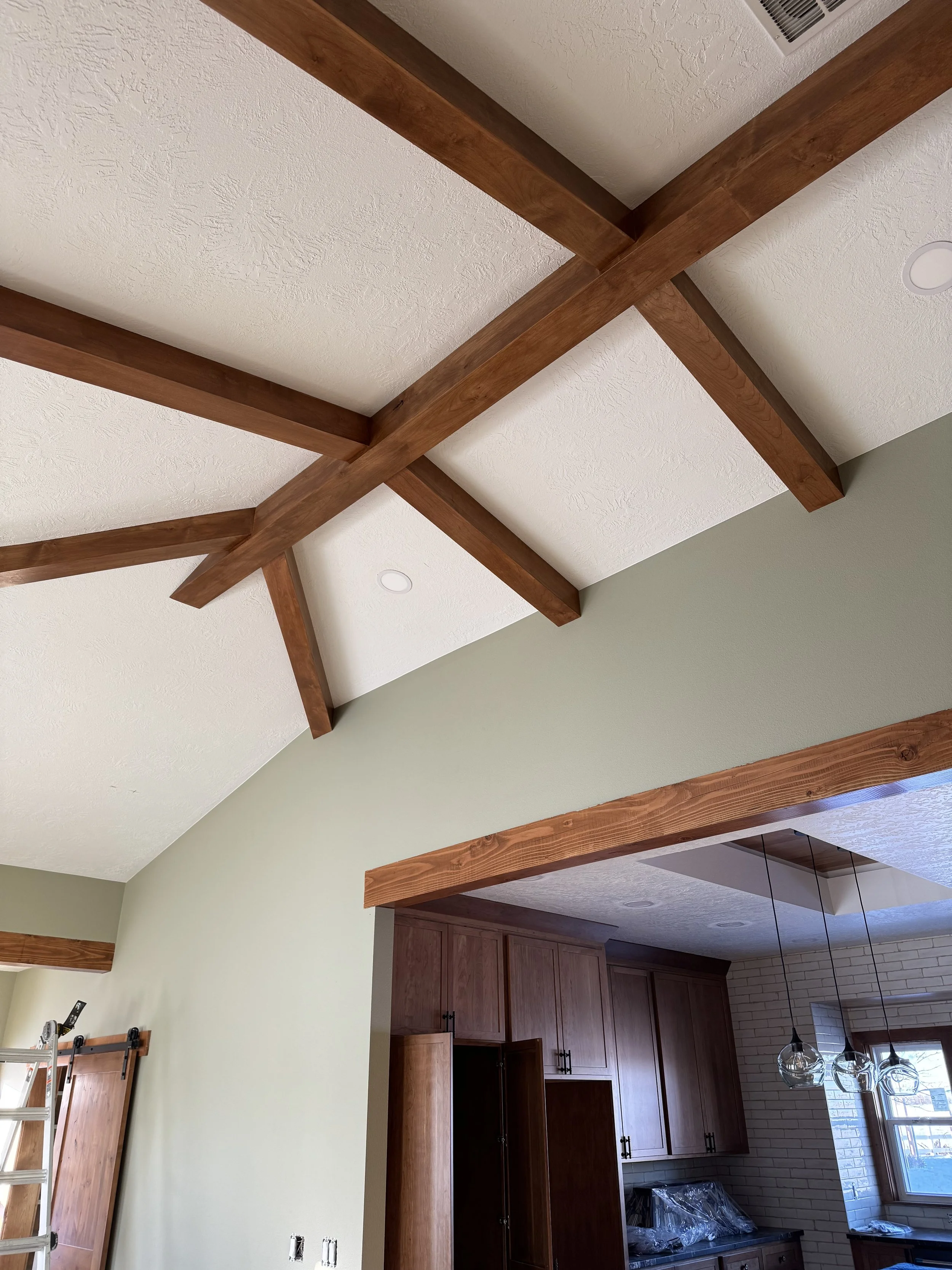 Interior view of a room with a vaulted ceiling featuring exposed wooden beams, light green painted walls, and a visible kitchen area with wooden cabinets, a window, and hanging light fixtures.