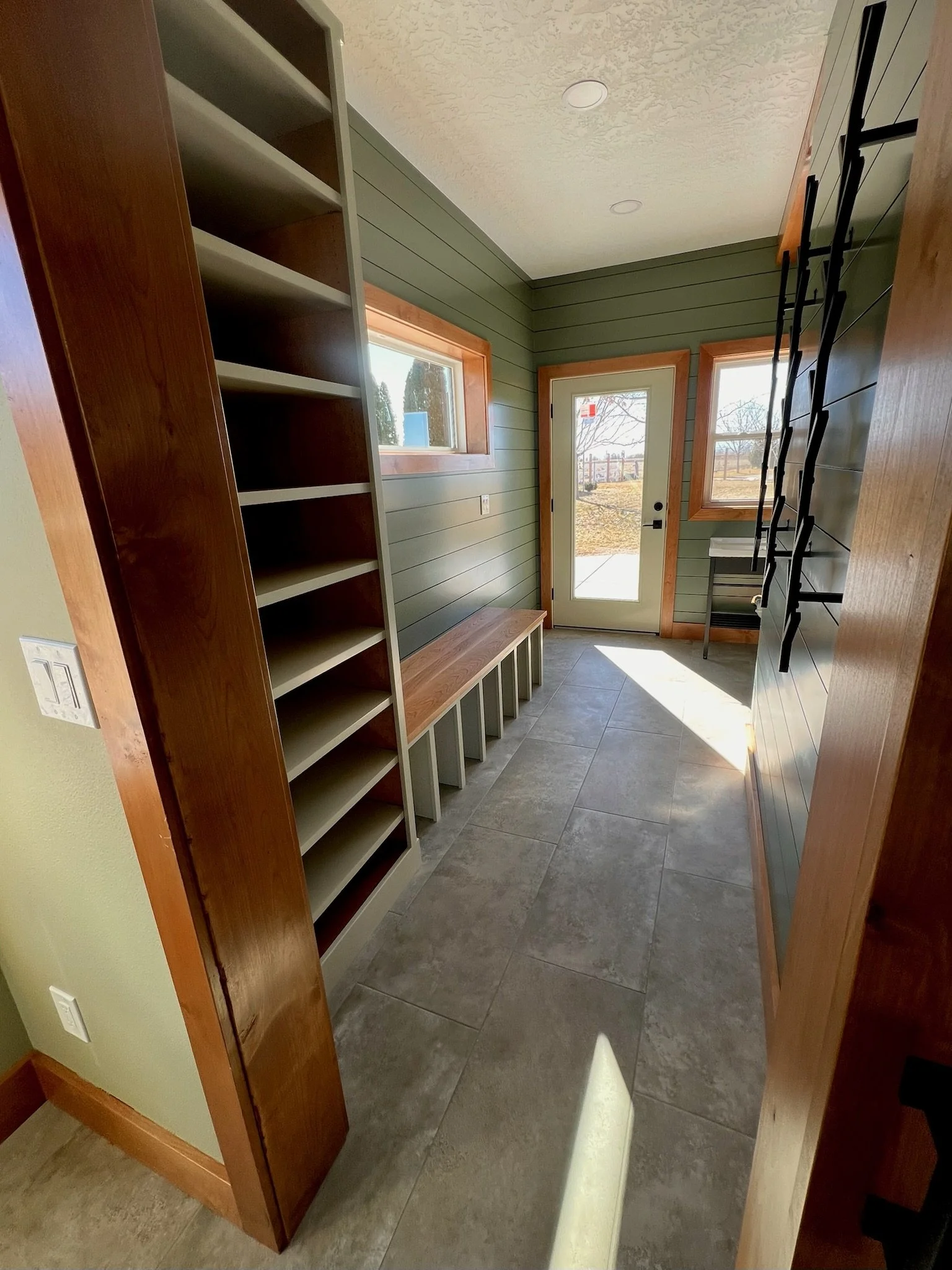 A small entryway with green wooden walls, a bench with cubbies underneath, and a tall built-in bookshelf. There's a door and two windows letting in natural light, with tile flooring.