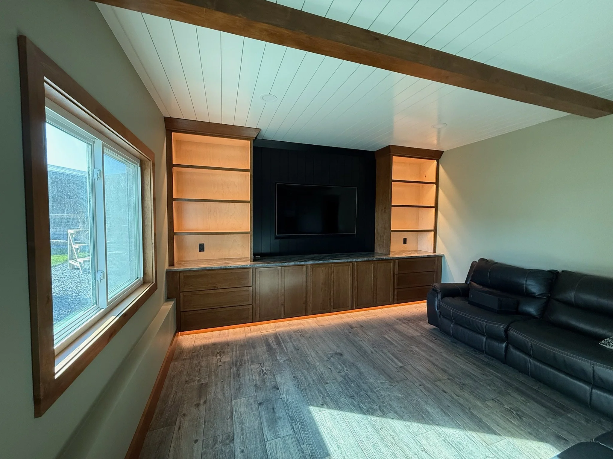 Living room with a large window, wooden built-in cabinets under a TV, and a black leather sofa.