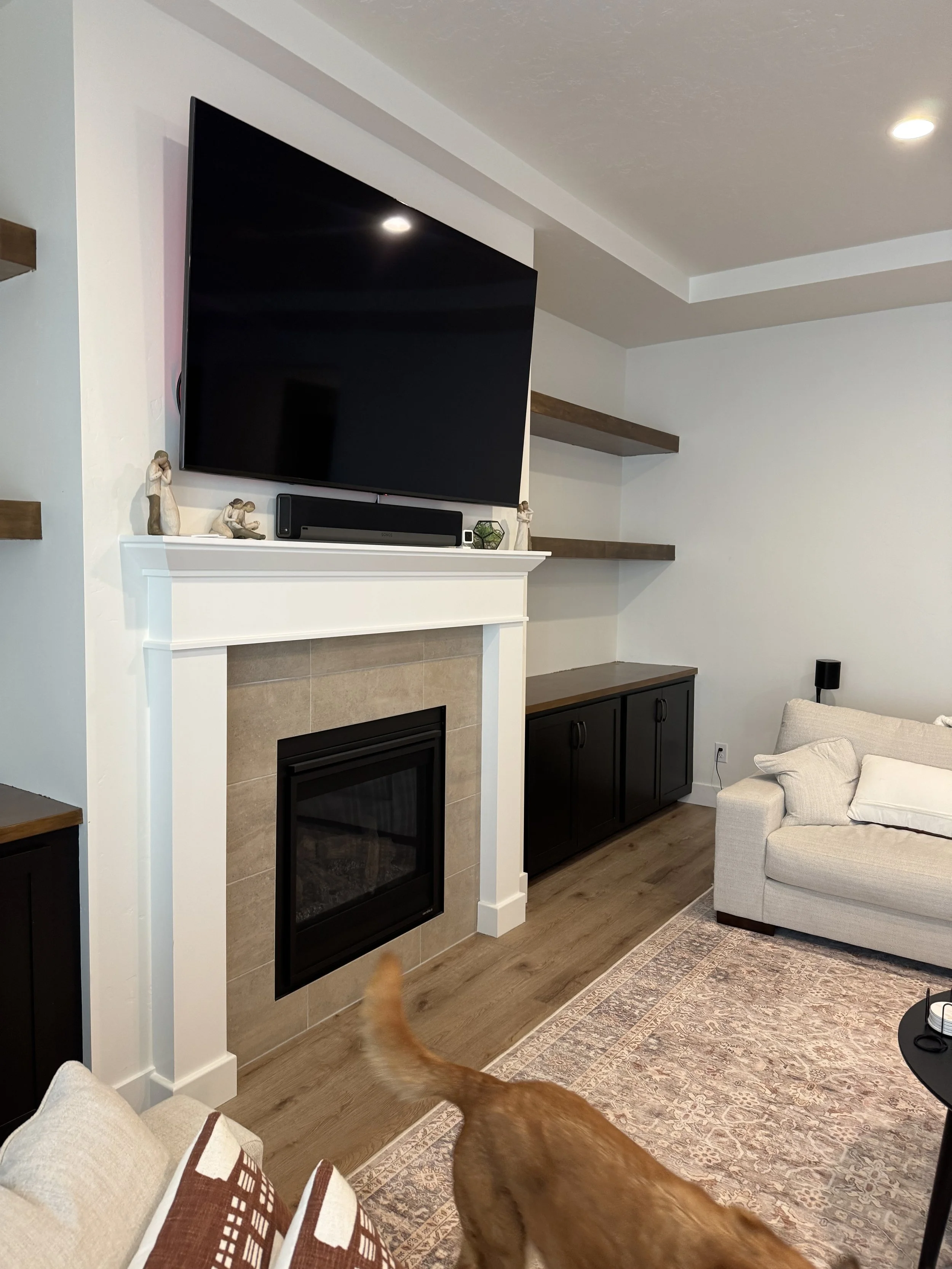 Living room with a mounted flat-screen TV above a fireplace with beige tiles, decorative figurines on the mantel, and built-in shelves on the wall. A corner of a white sofa with pillows and a dog with a raised tail are visible.
