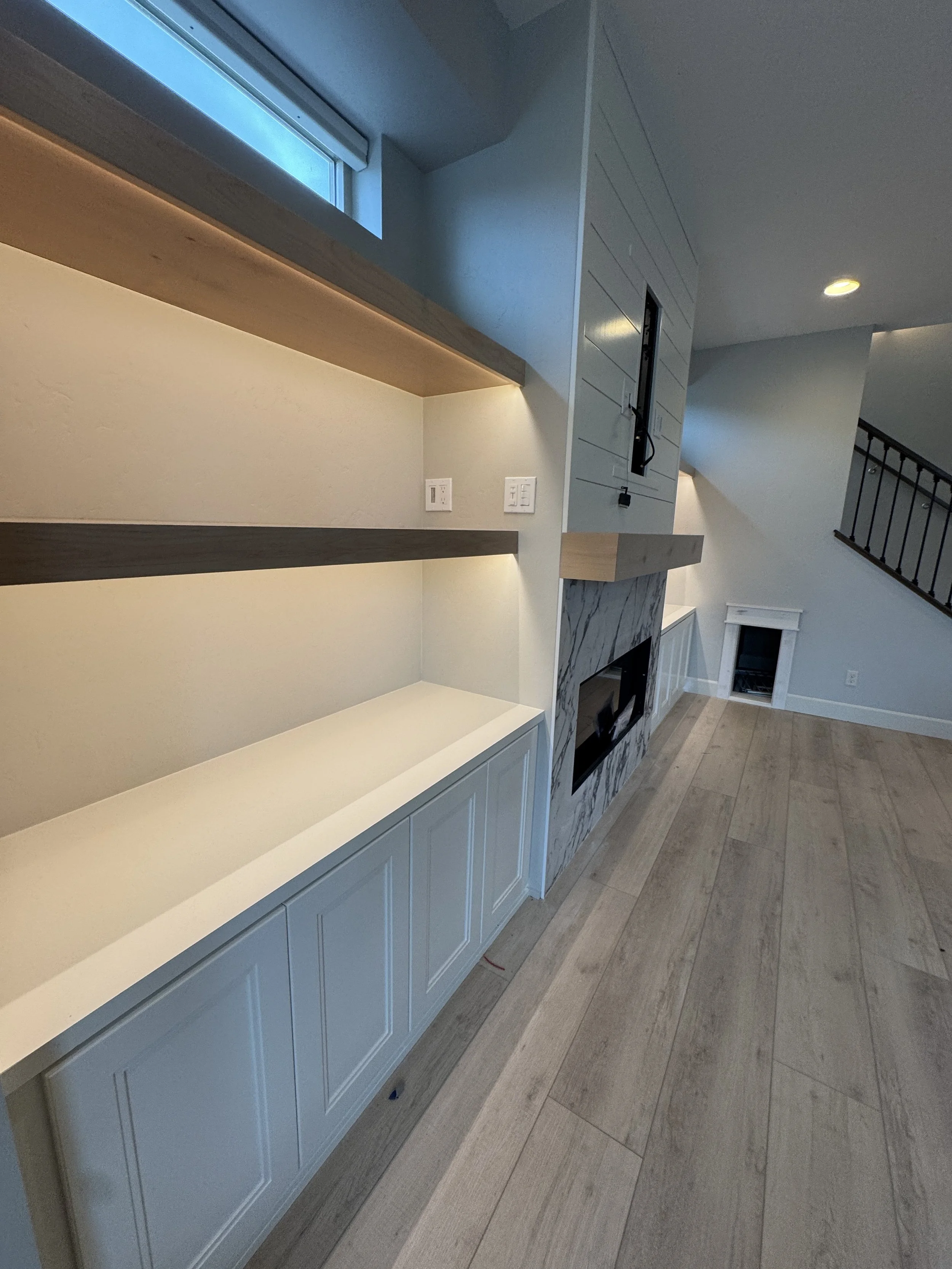 Interior view of a living room with built-in shelves, a fireplace with a marble surround, hardwood flooring, and a staircase.