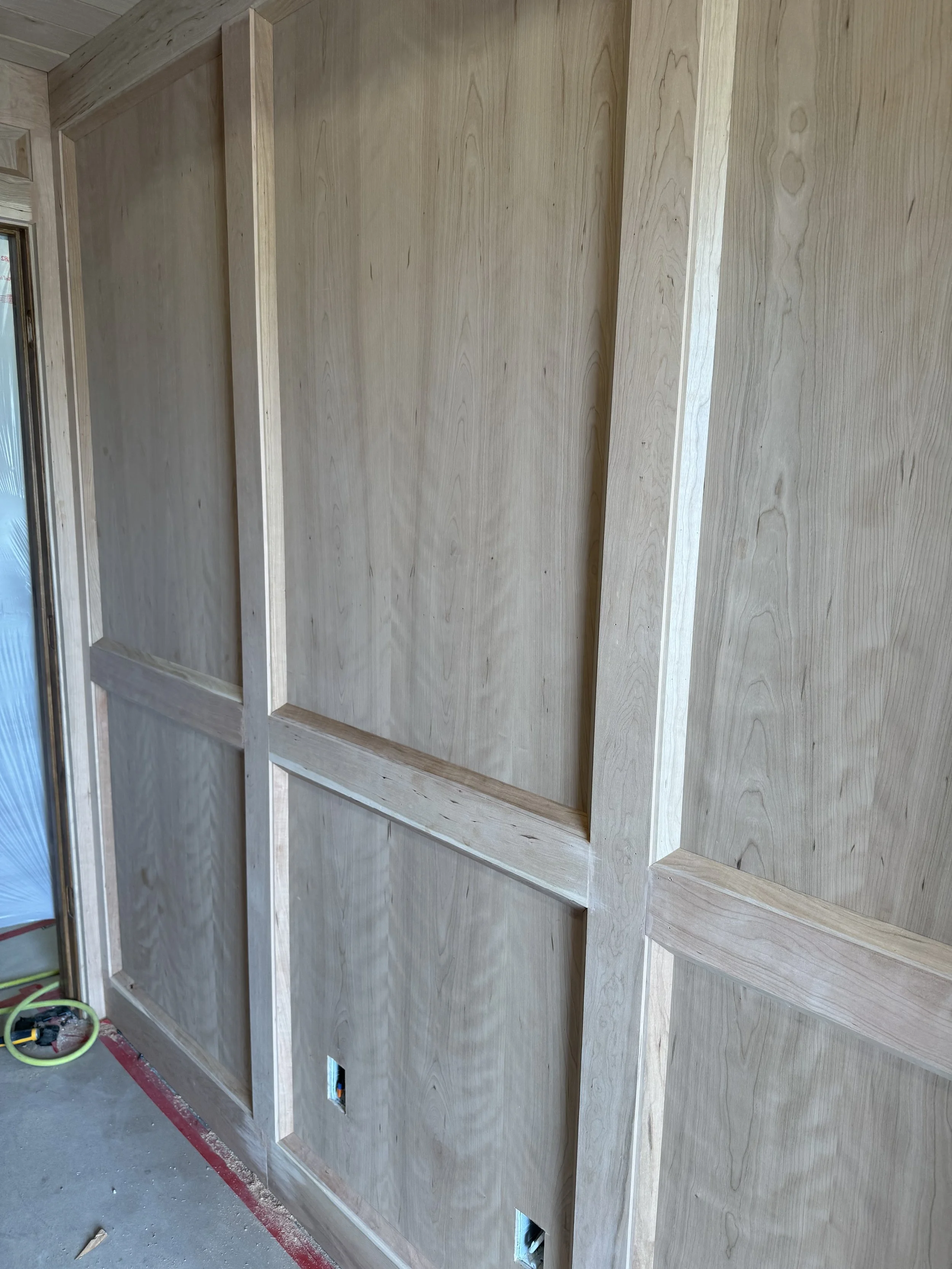 Wooden wall framing with plywood panels installed, electrical outlets and wiring access holes at the bottom, construction tools on the floor, near a sliding glass door.