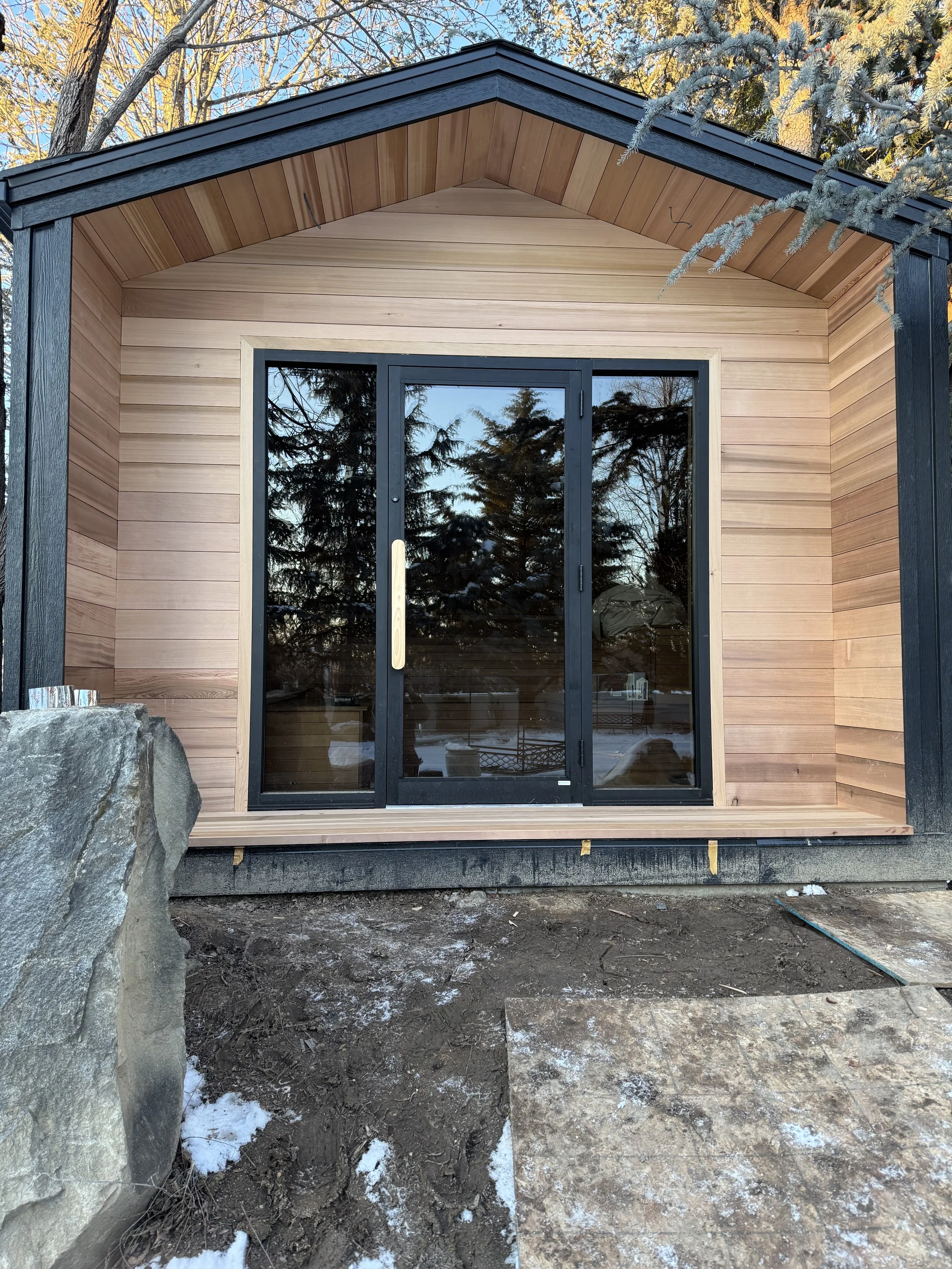 Newly constructed small wooden cabin with sliding glass doors, surrounded by outdoor trees and partially covered in snow.