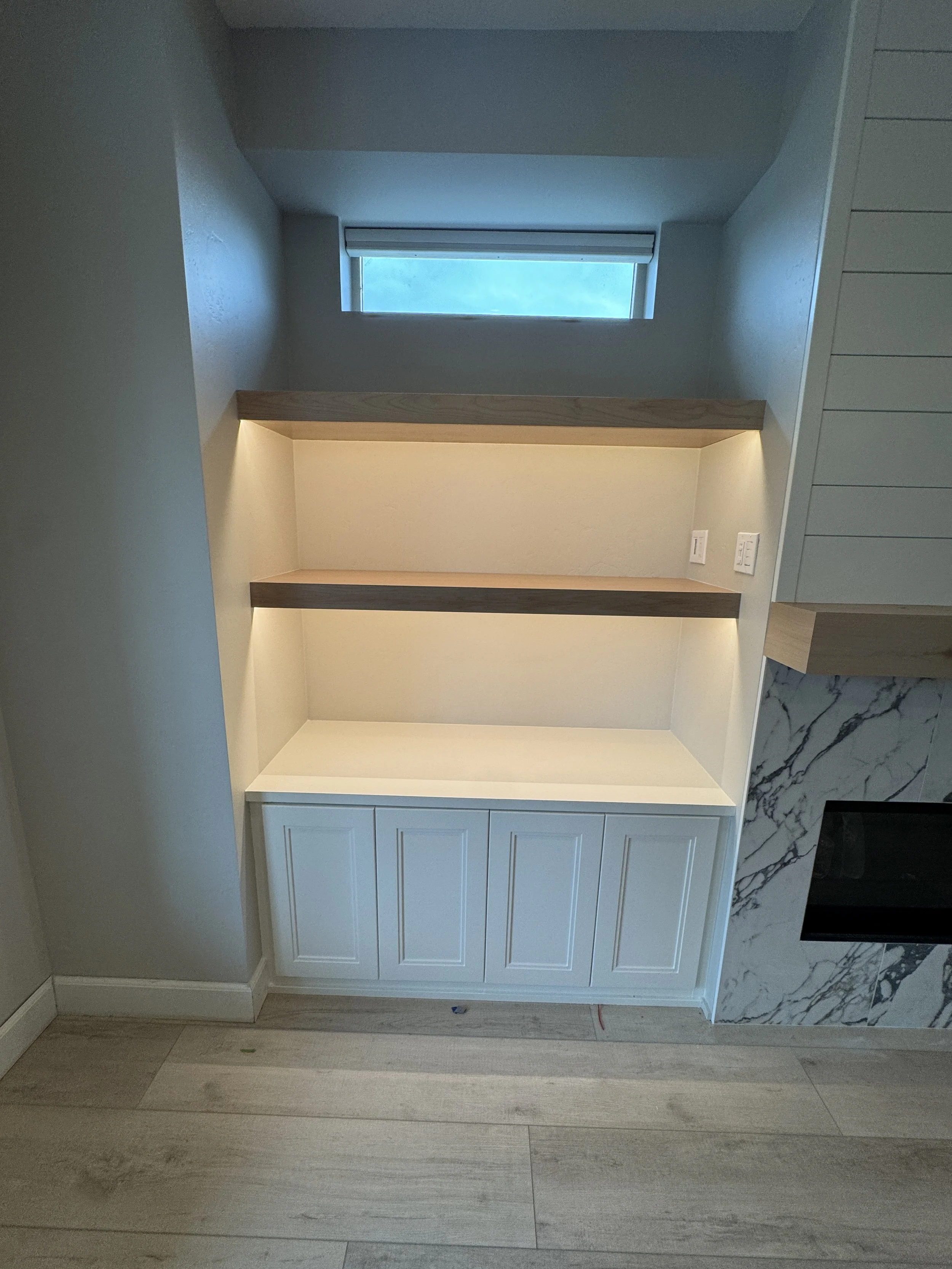 Empty built-in shelving with two wooden shelves, LED lighting, electrical outlets, and cabinets at the bottom, next to a white marble fireplace and large window.
