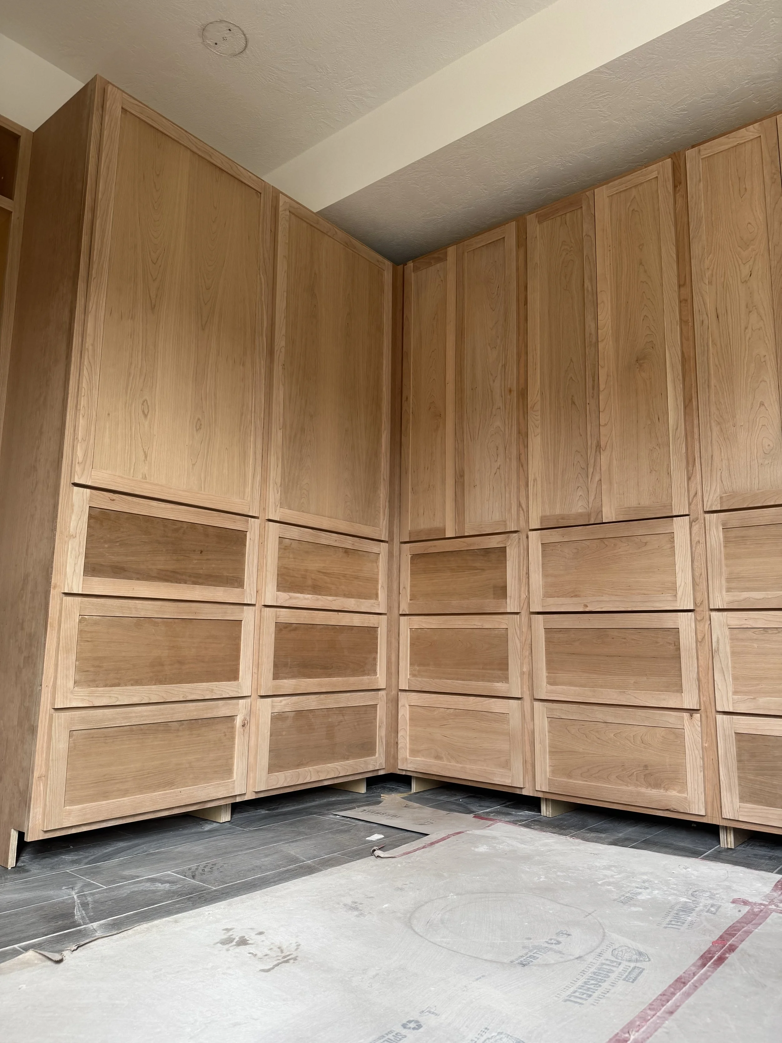 Unfinished wooden kitchen cabinets with open drawers, located in a corner of a room with a concrete floor and a white ceiling.