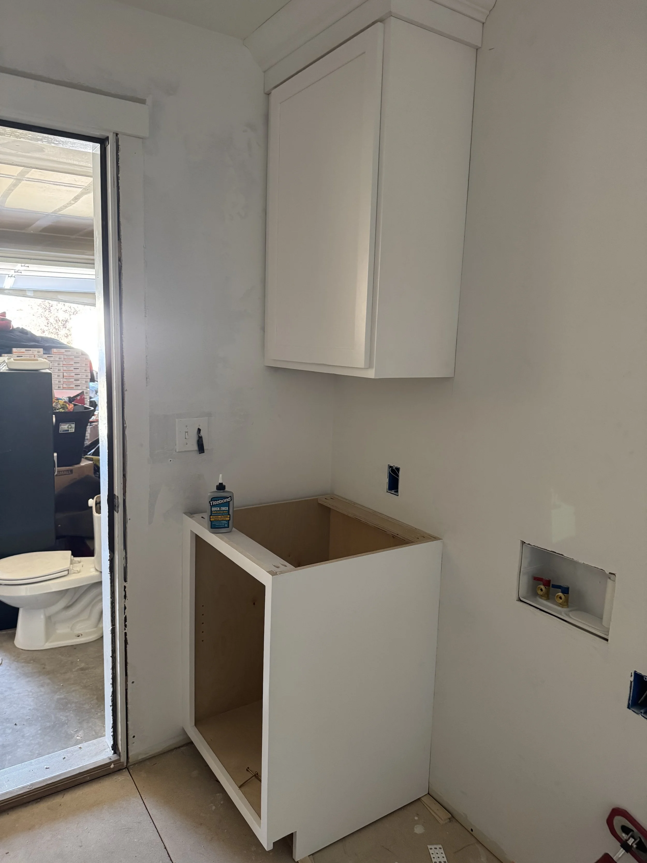 Laundry area under construction with a white cabinet above and a new washing machine or dryer space below, a bottle of glue on top, and plumbing connections on the wall.
