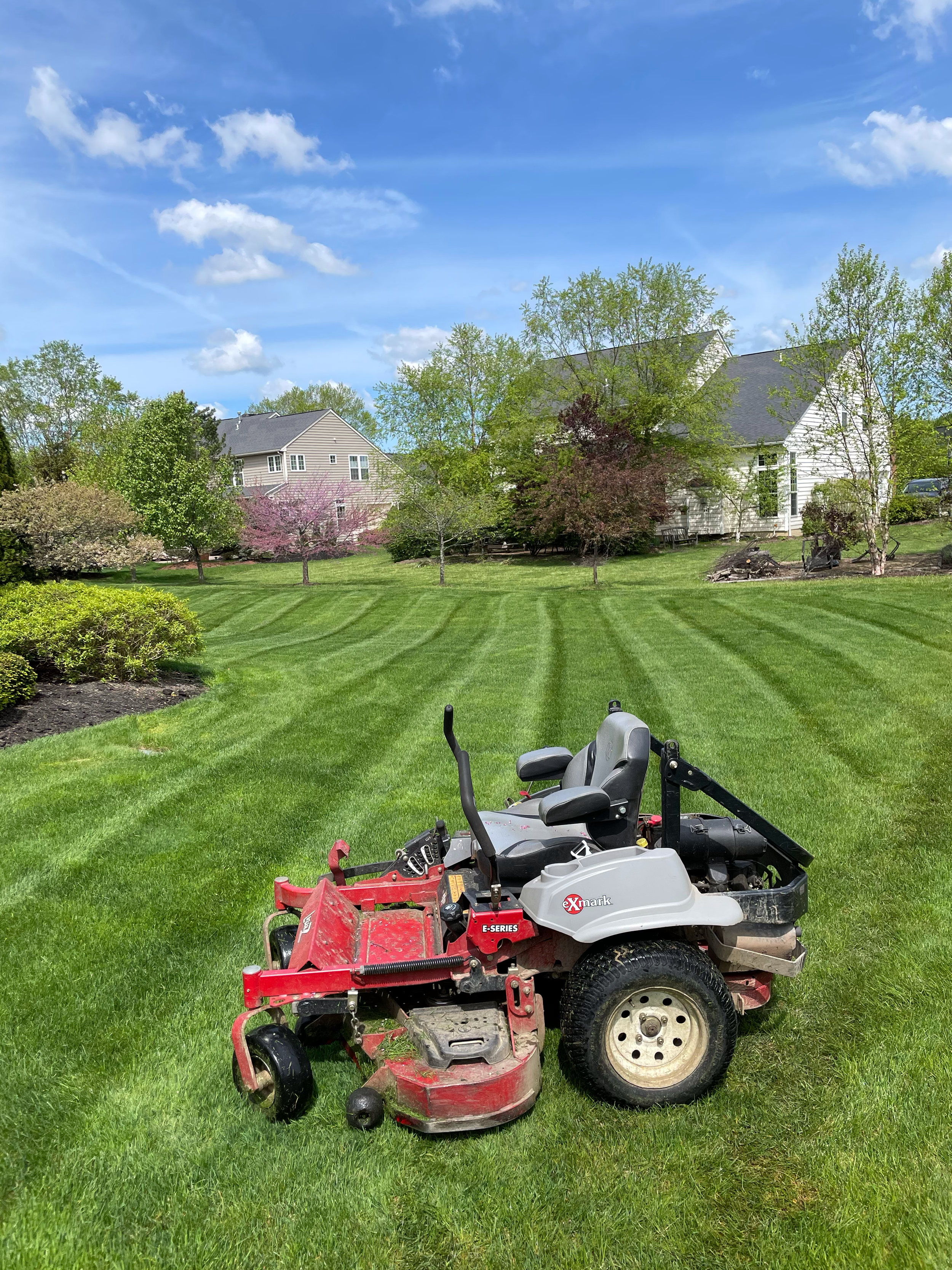 A red and gray riding mower on a well-manicured lawn with houses and trees in the background under a partly cloudy sky.