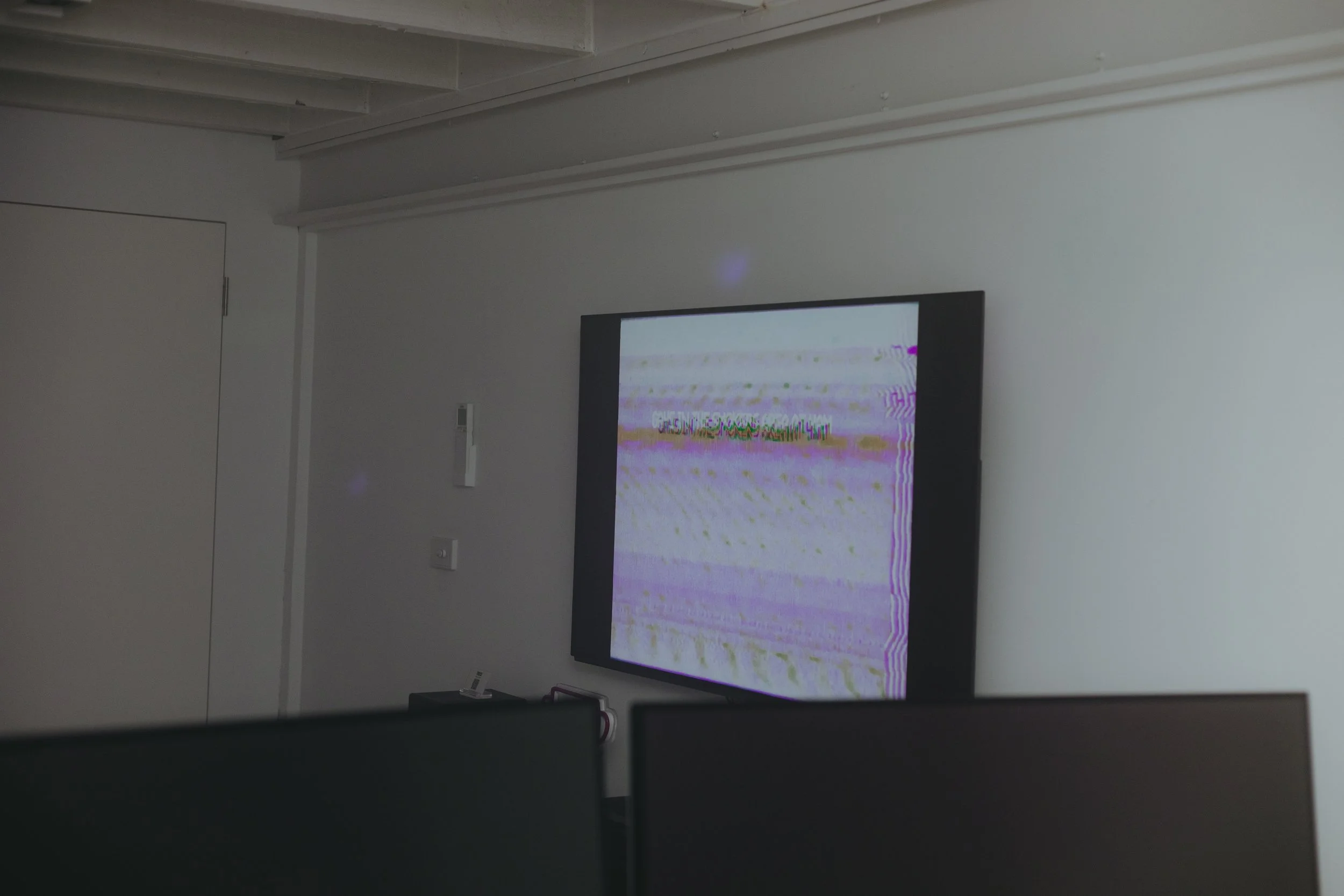 A flat-screen television displaying a distorted, glitchy image with colorful lines and static, mounted on a plain white wall in a modern room with minimal decor.