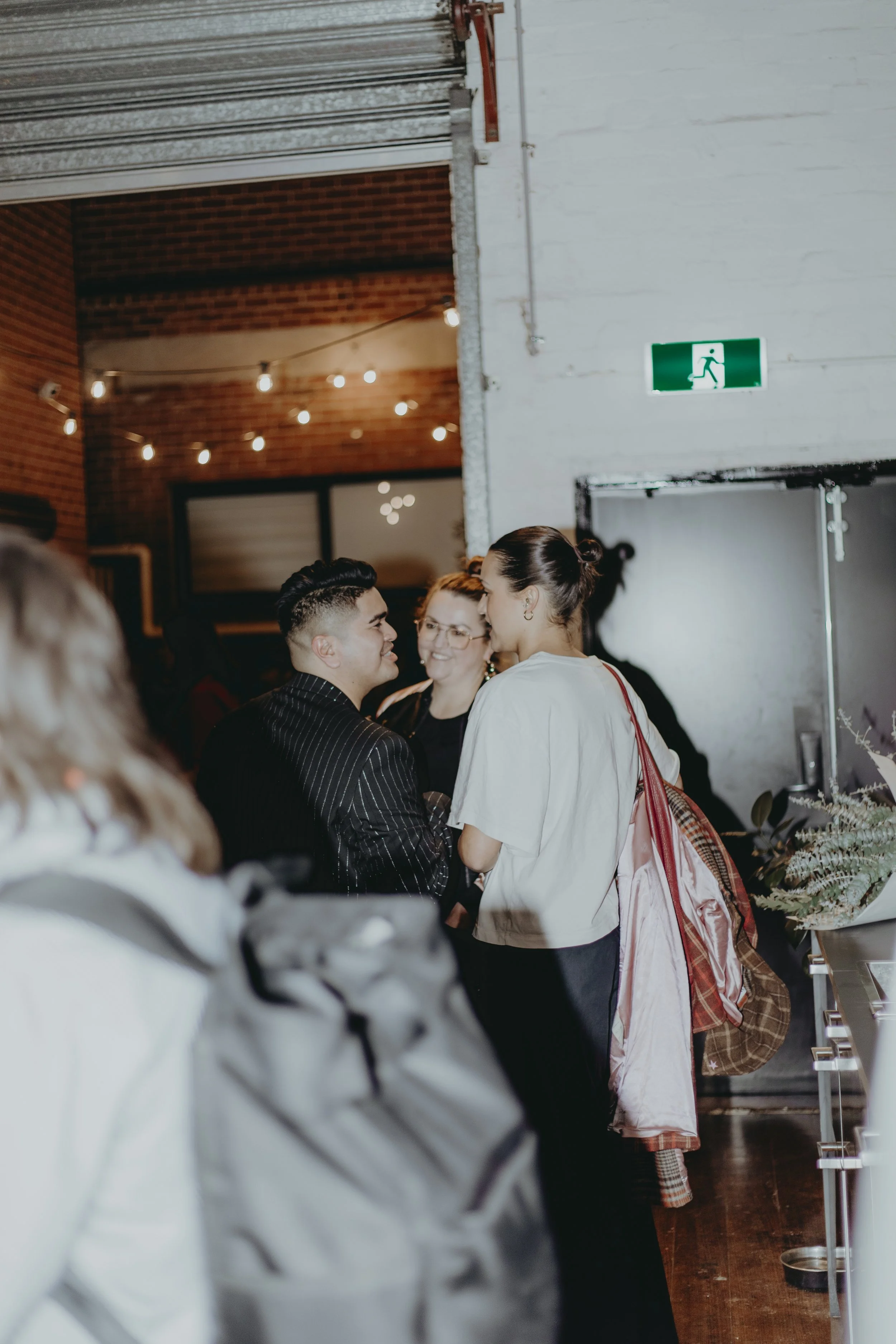 A group of people standing indoors near a white wall with an emergency exit sign. The room has string lights hanging from the ceiling and a brick wall in the background. The floor appears to be wooden, and there are some plants and decorations visibl