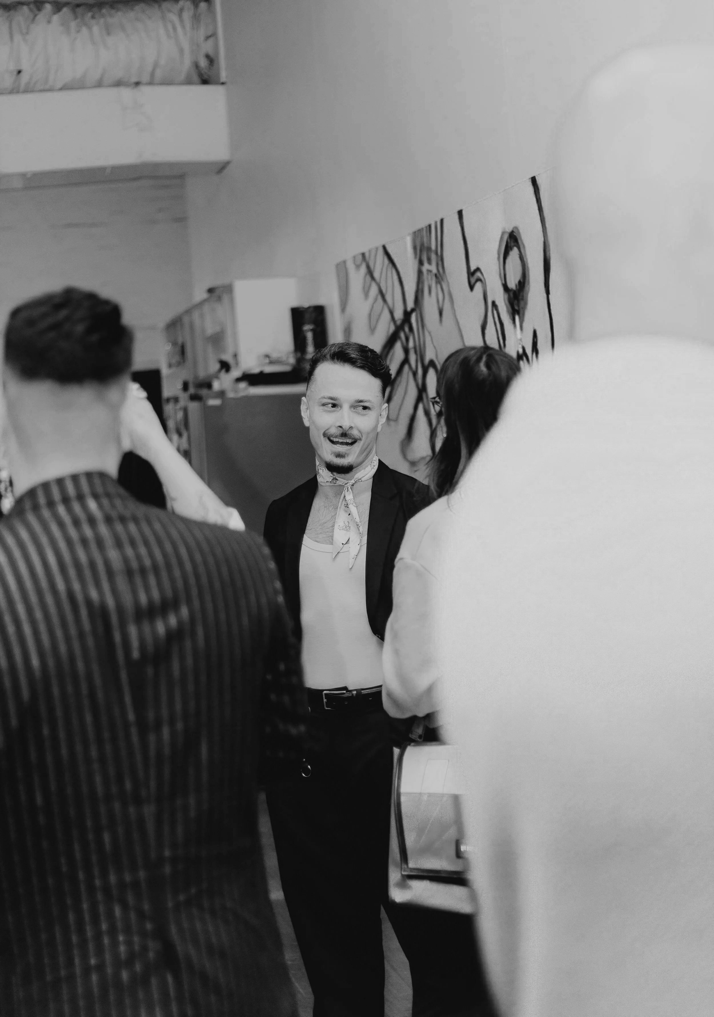 A man with a mustache and beard in a black suit and white shirt, smiling and talking to several people in a hallway decorated with abstract black graffiti on the wall.