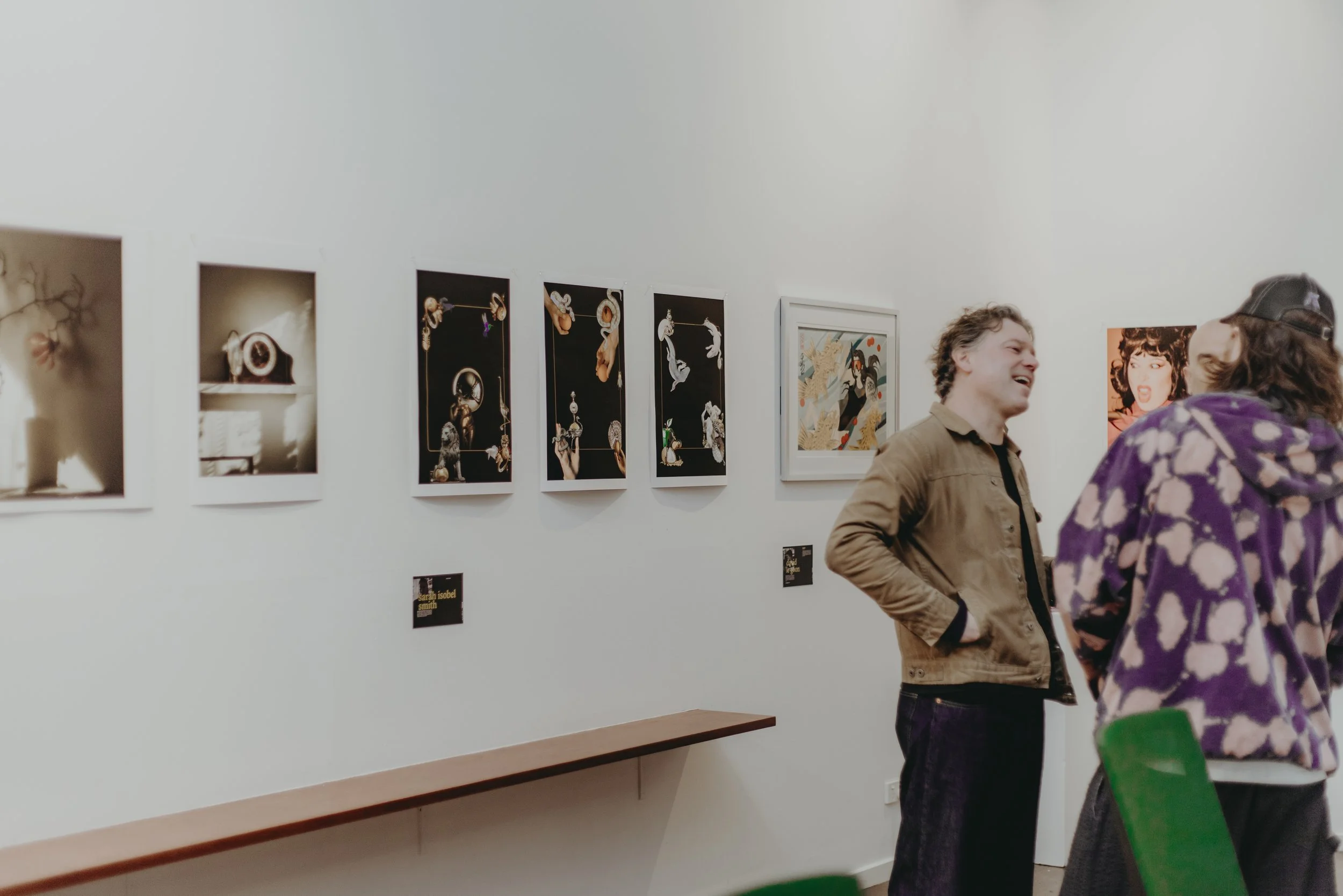 People are walking through an art gallery with colorful artwork and photographs displayed on the white wall.