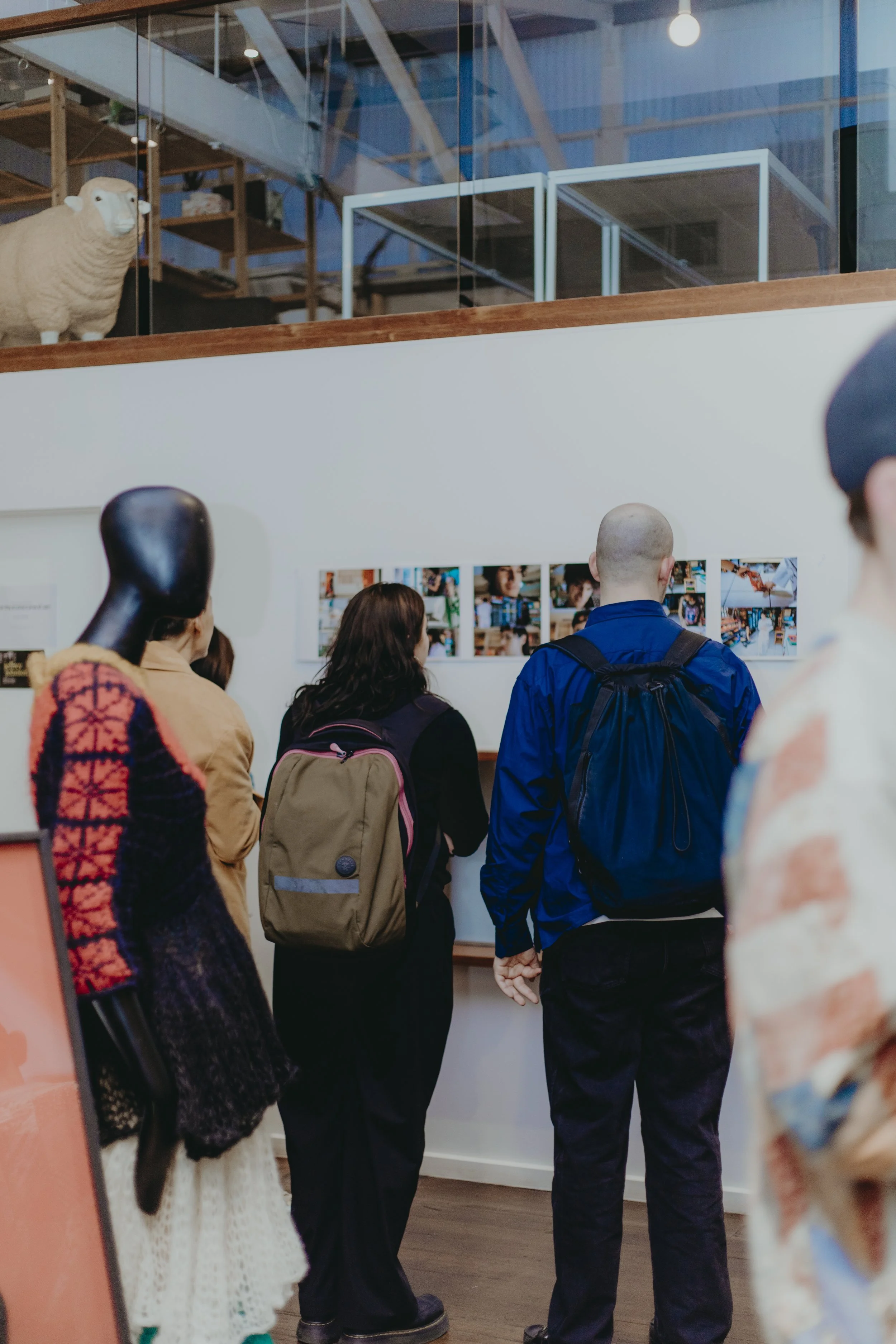 People standing in line at an indoor art gallery or exhibition, looking at photographs on the wall. An animal sculpture of a sheep is visible on the upper level behind a glass barrier.