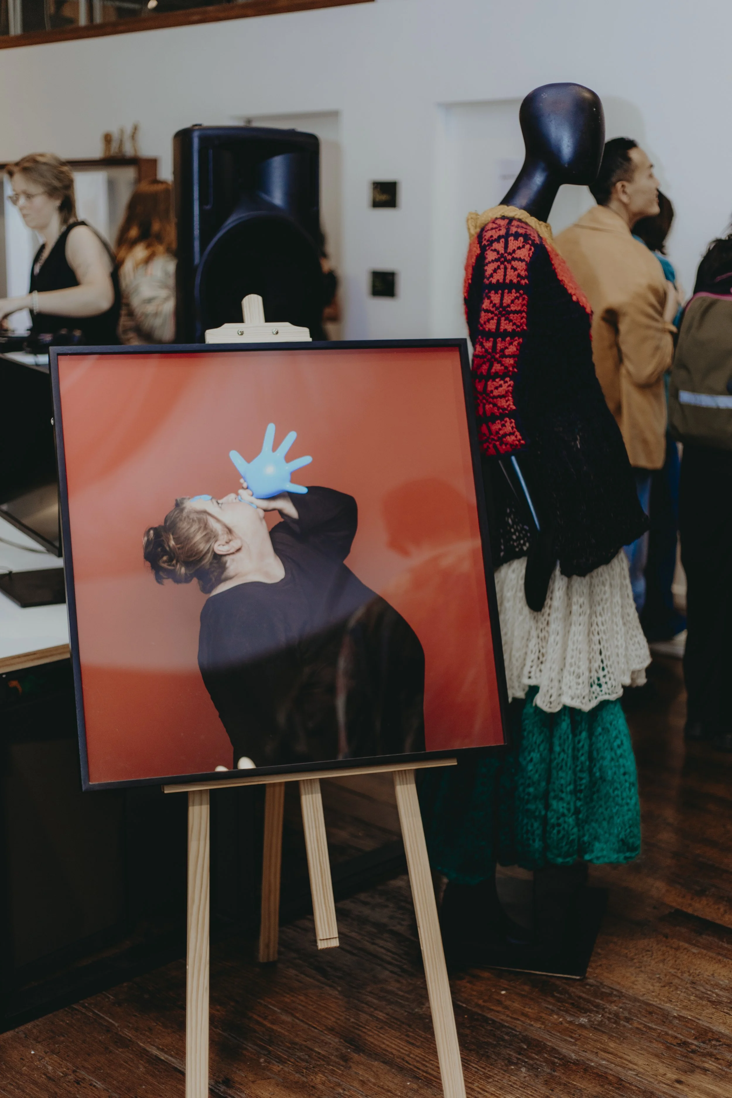 People in an art gallery observing artwork and a portrait on an easel, with a woman dressed in period costume and a woman in a black top on the left, and a woman with a yellow coat and long skirt on the right.