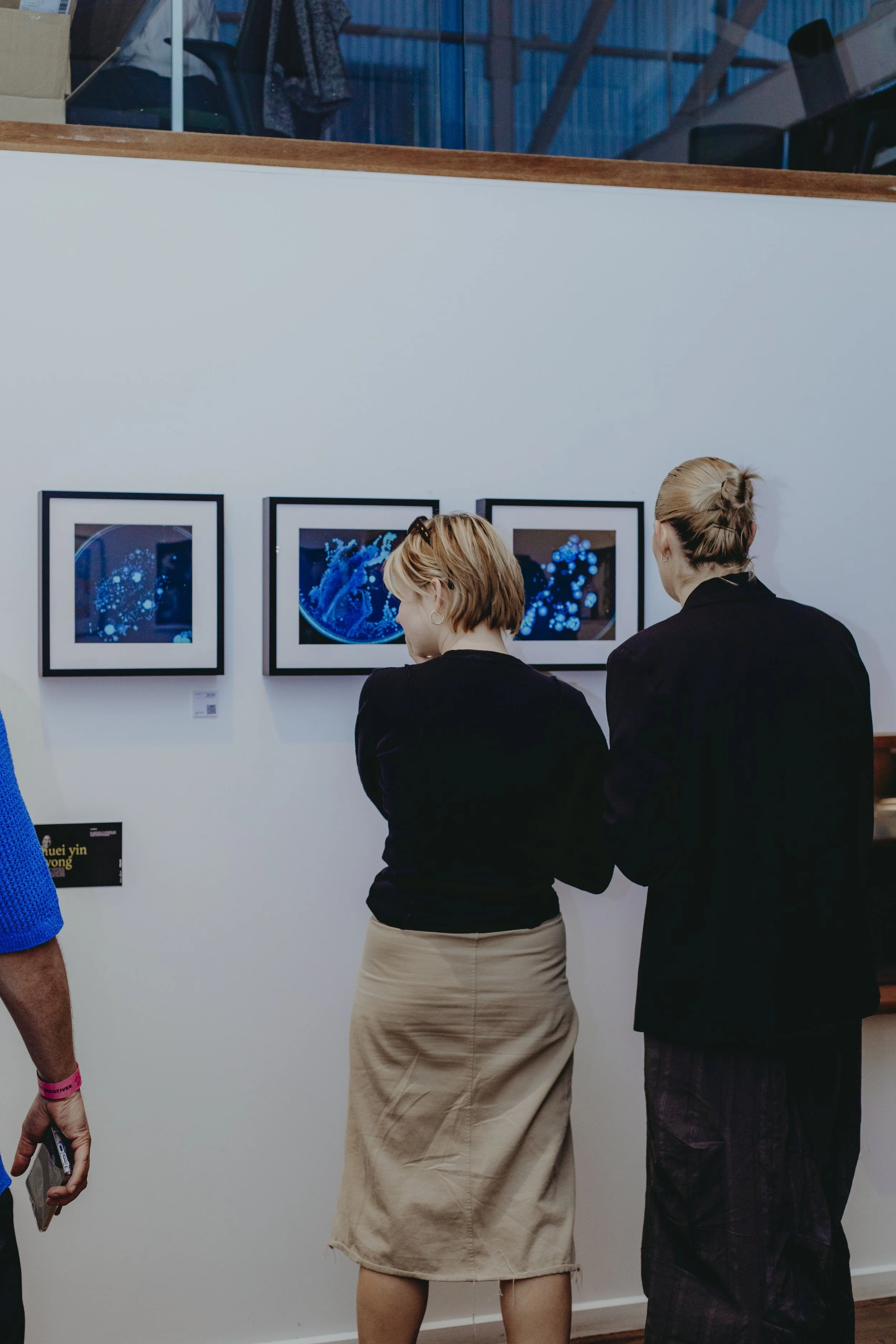 Two women are viewing framed displayed digital artwork featuring blue light patterns at an art gallery. One woman is wearing a beige skirt and the other is dressed in black. Other people and artworks are visible in the background.