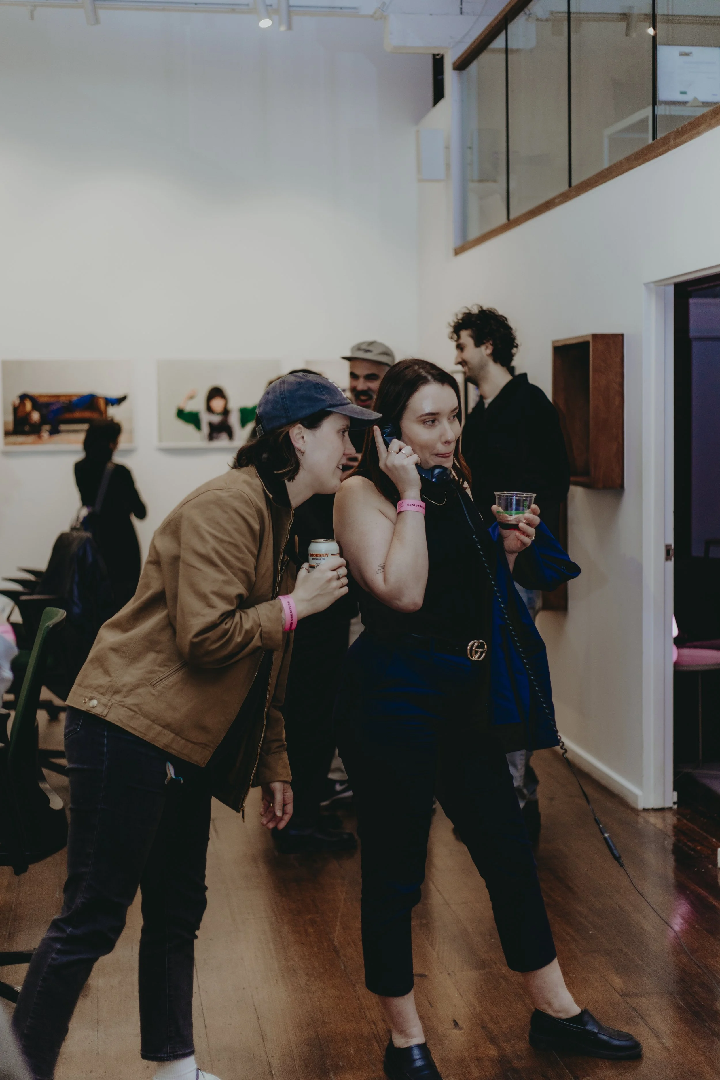 Several people in an art gallery, some engaged in conversation, others looking at artwork on the walls. Women are wearing casual clothing, including a woman with a phone and drinks, and a man in a blazer. The gallery features paintings on the white w