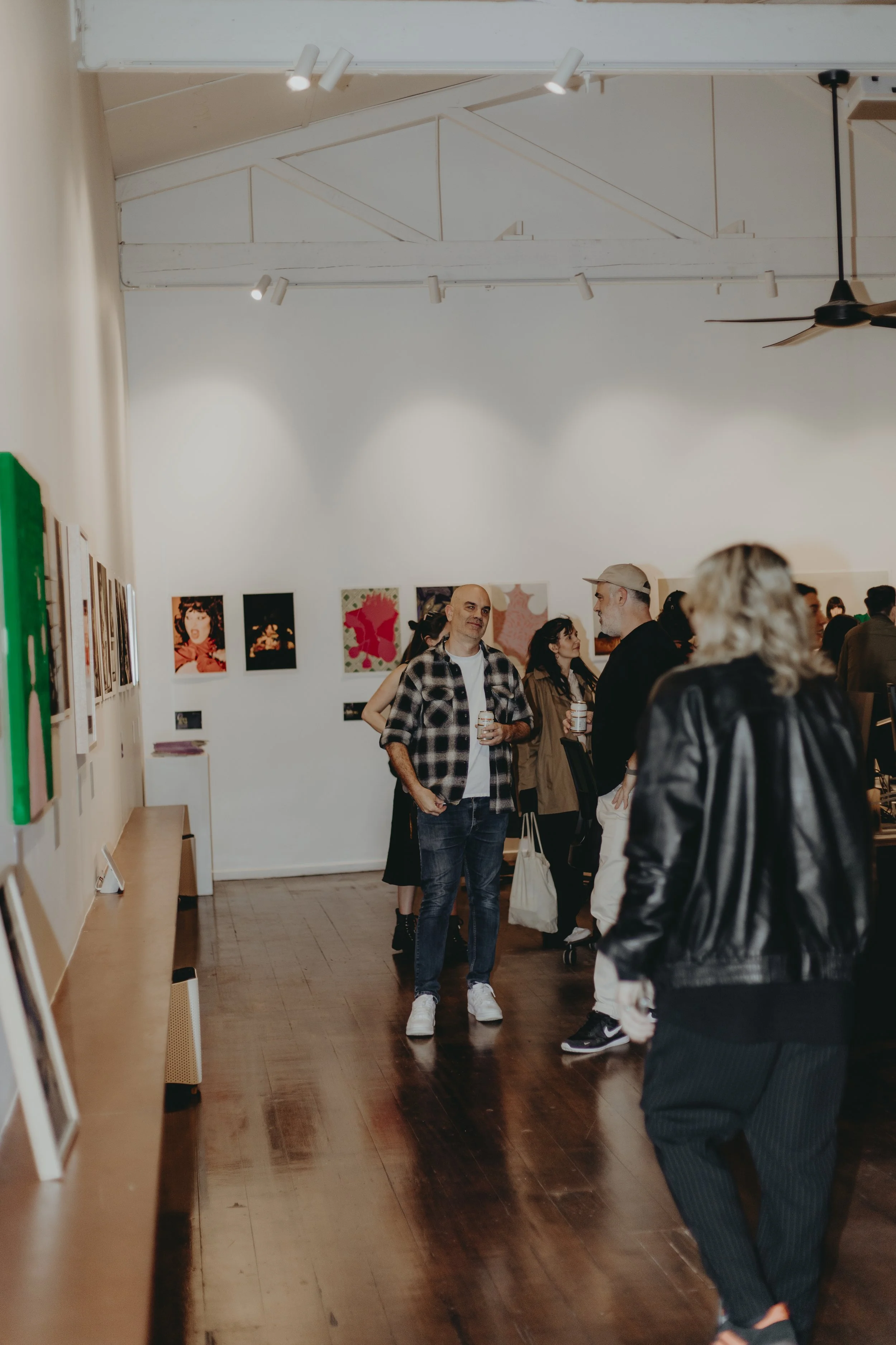 People attending an art gallery exhibition, viewing and conversing in a brightly lit room with white walls and wooden floors.