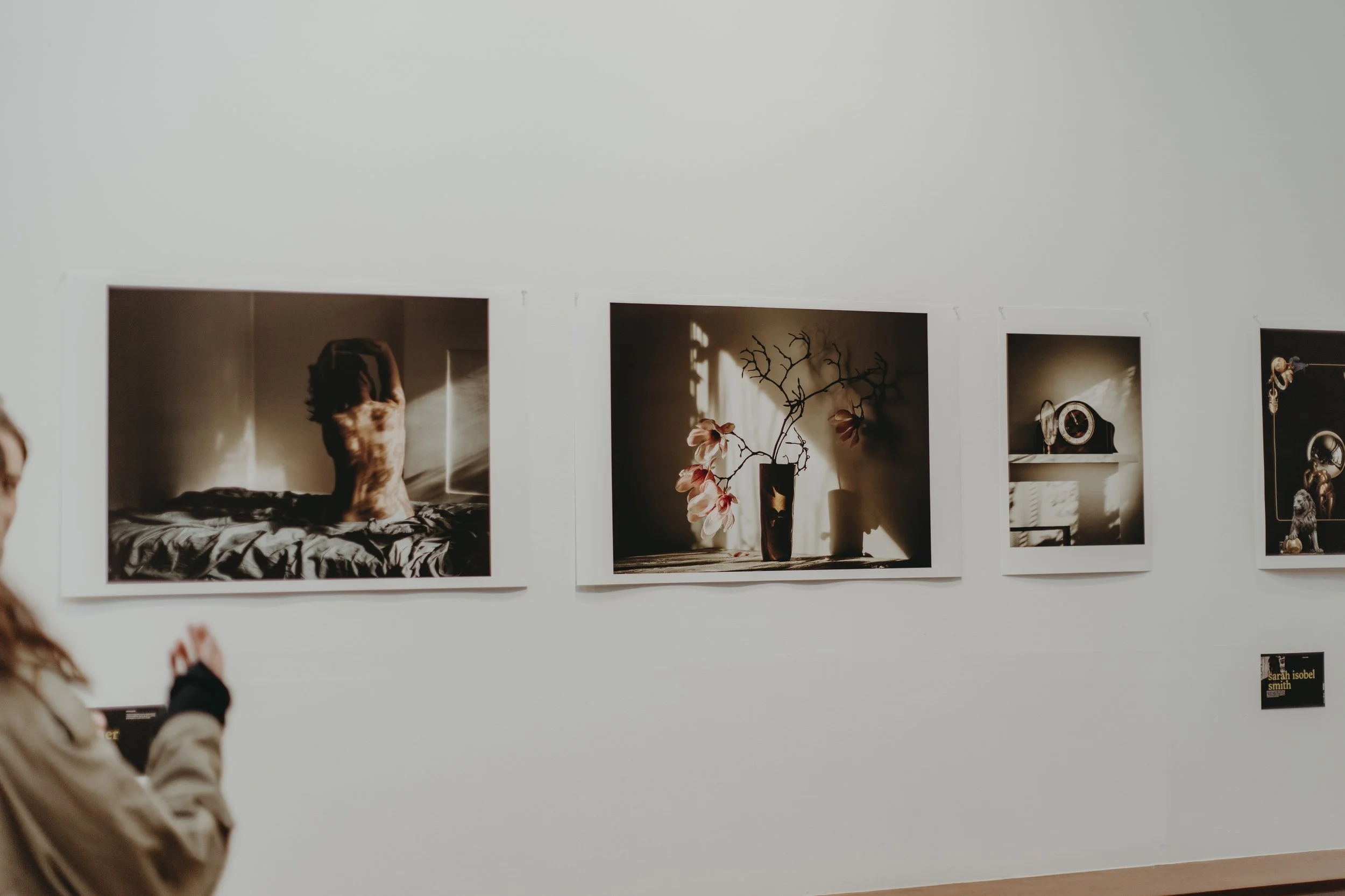 Photographic art exhibit on a gallery wall featuring three framed images: a sculpture of a person with hands covering their face, a flower vase with pink blossoms and shadows cast on the wall, and a vintage clock with decorative objects. A woman in t