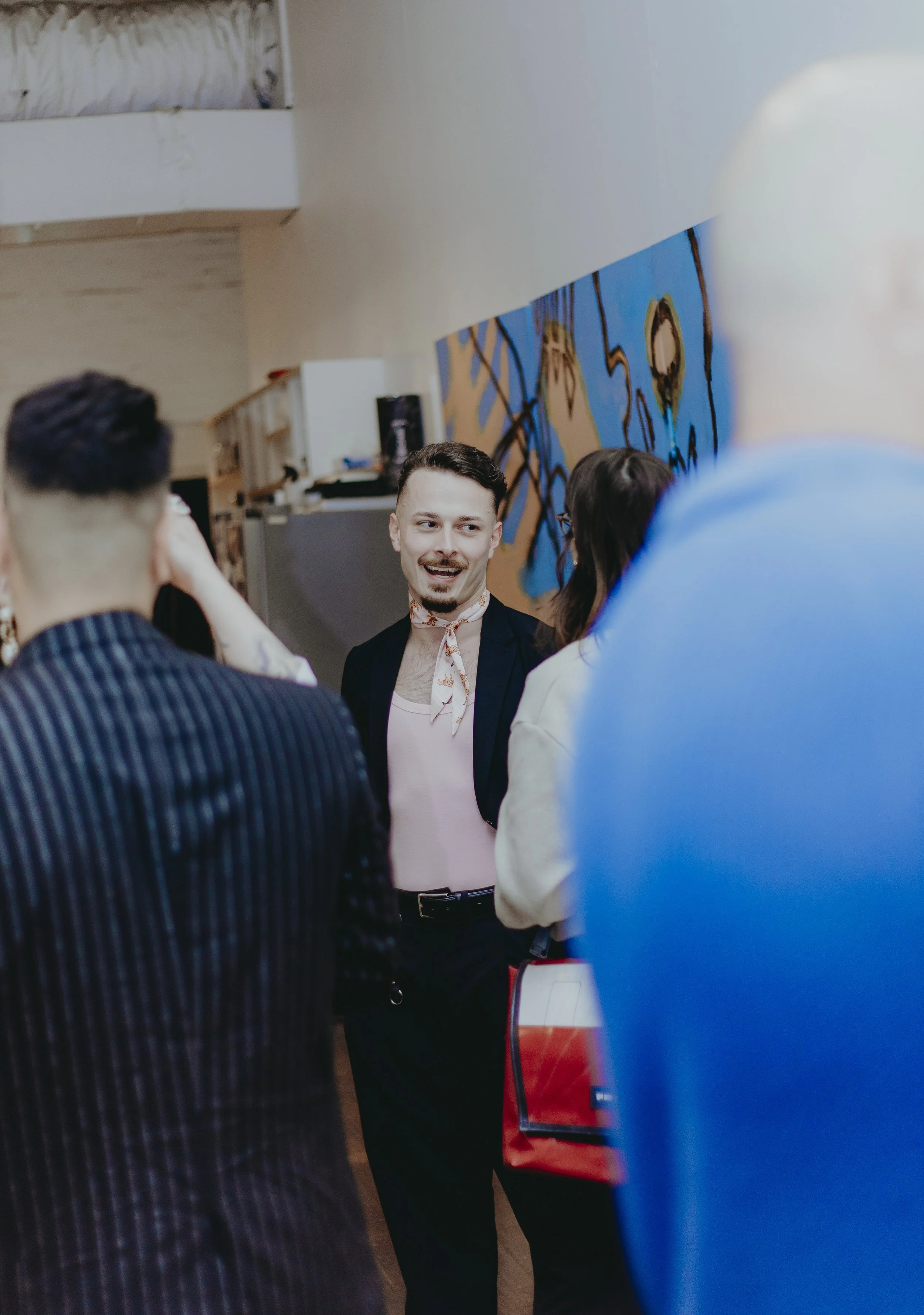 A man with a mustache and goatee, wearing a white shirt with a floral tie and a black jacket, is standing in a room with people around him. The background features a wall with abstract art in blue, beige, and black colors. The setting appears to be i