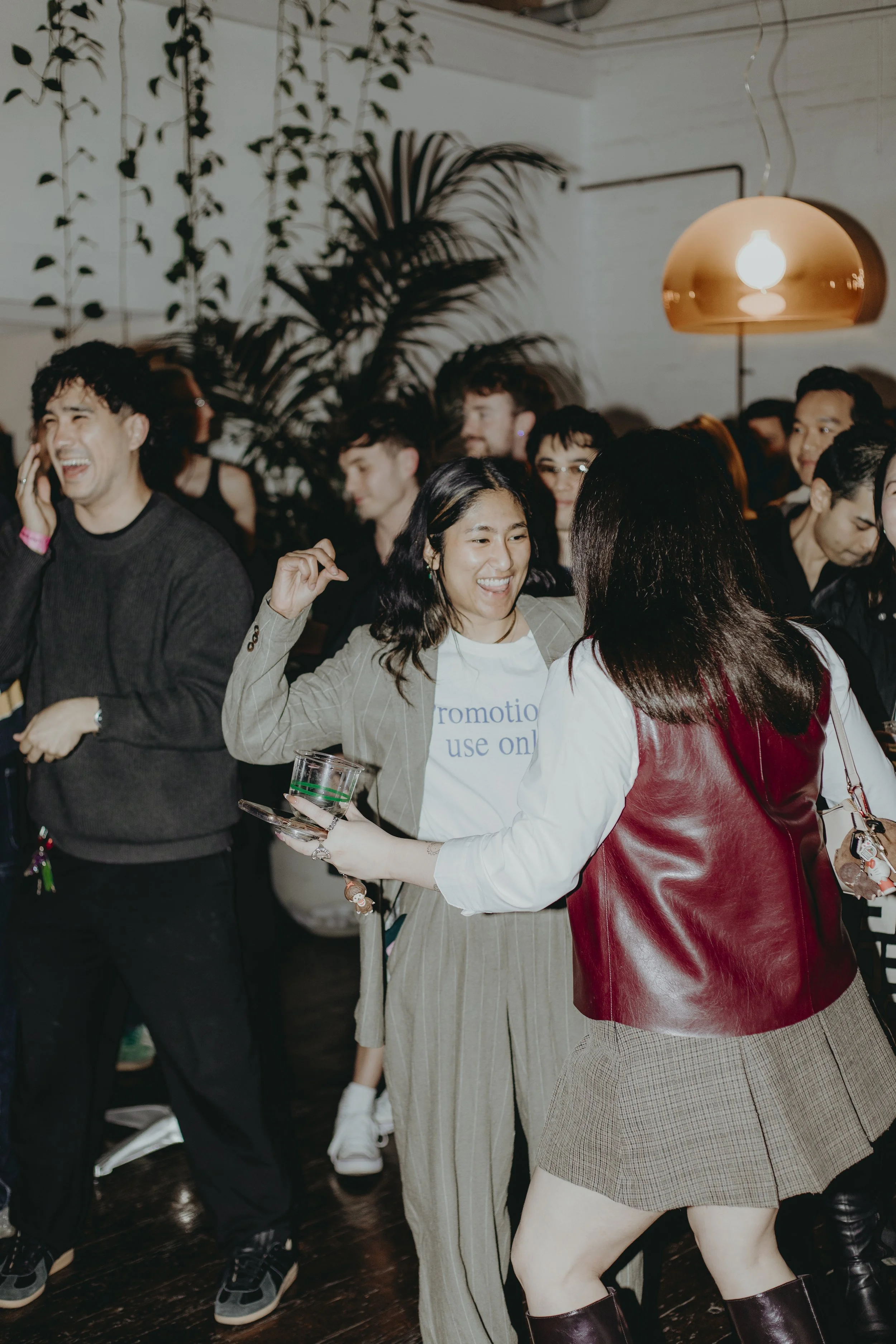 People dancing and socializing at a party in a room with white walls and a hanging copper-colored lamp. Smiling and enjoying themselves.